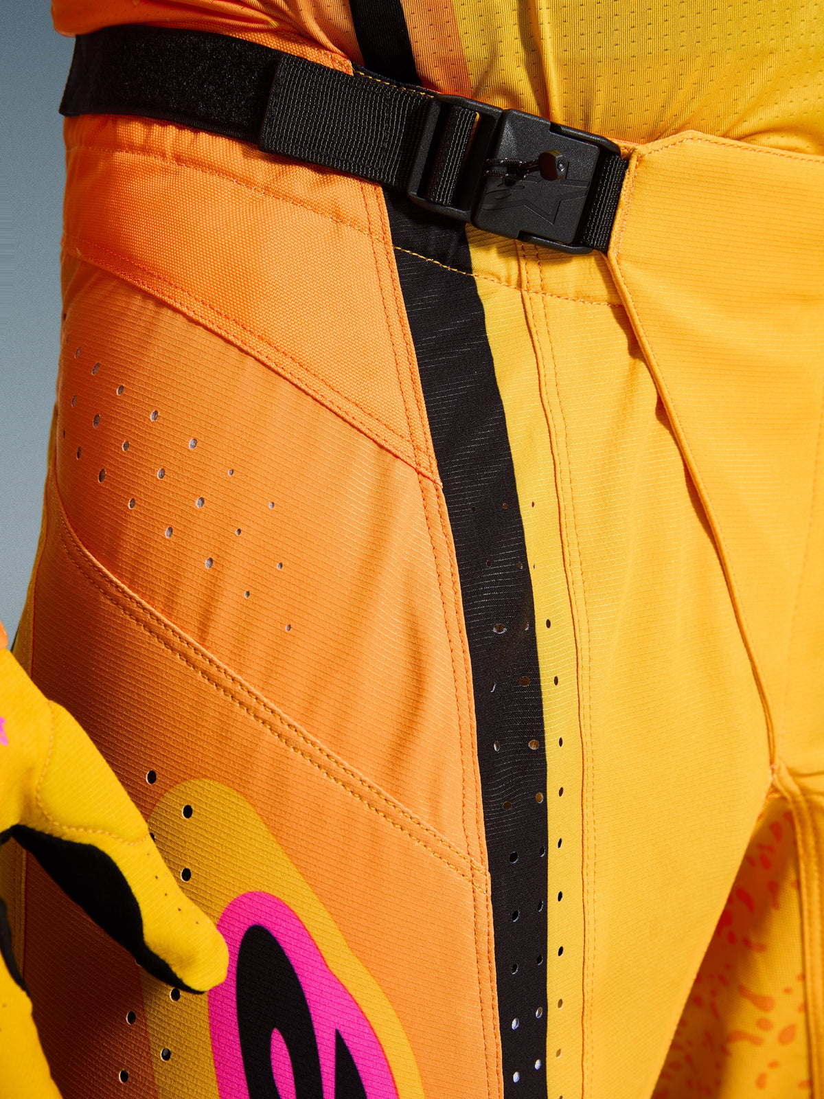 Alpinestars 2026 Techstar Nomur Pants, pantalones de motocross, naranja, amarillo y rosa, primer plano de la zona de la cintura con el cierre de hebilla magnética SX2, correa de ajuste negra y perforaciones de ventilación cortadas con láser en material ripstop duradero
