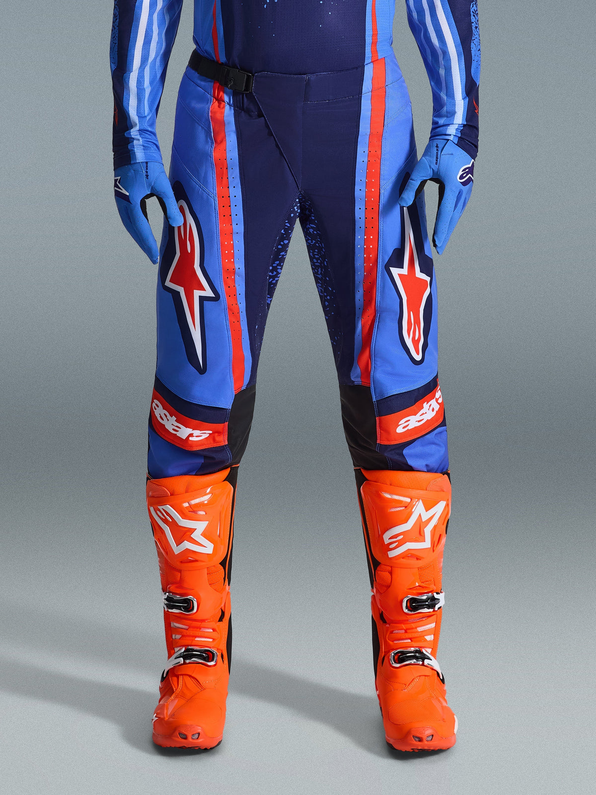 Alpinestars 2026 Techstar Nomur Pants, Motocross Pants, Dark Navy, Orange, and UCLA Blue, featuring bold orange stripes and large white star logos on the thighs, worn with bright orange racing boots and blue gloves, athletic race fit for off-road performance