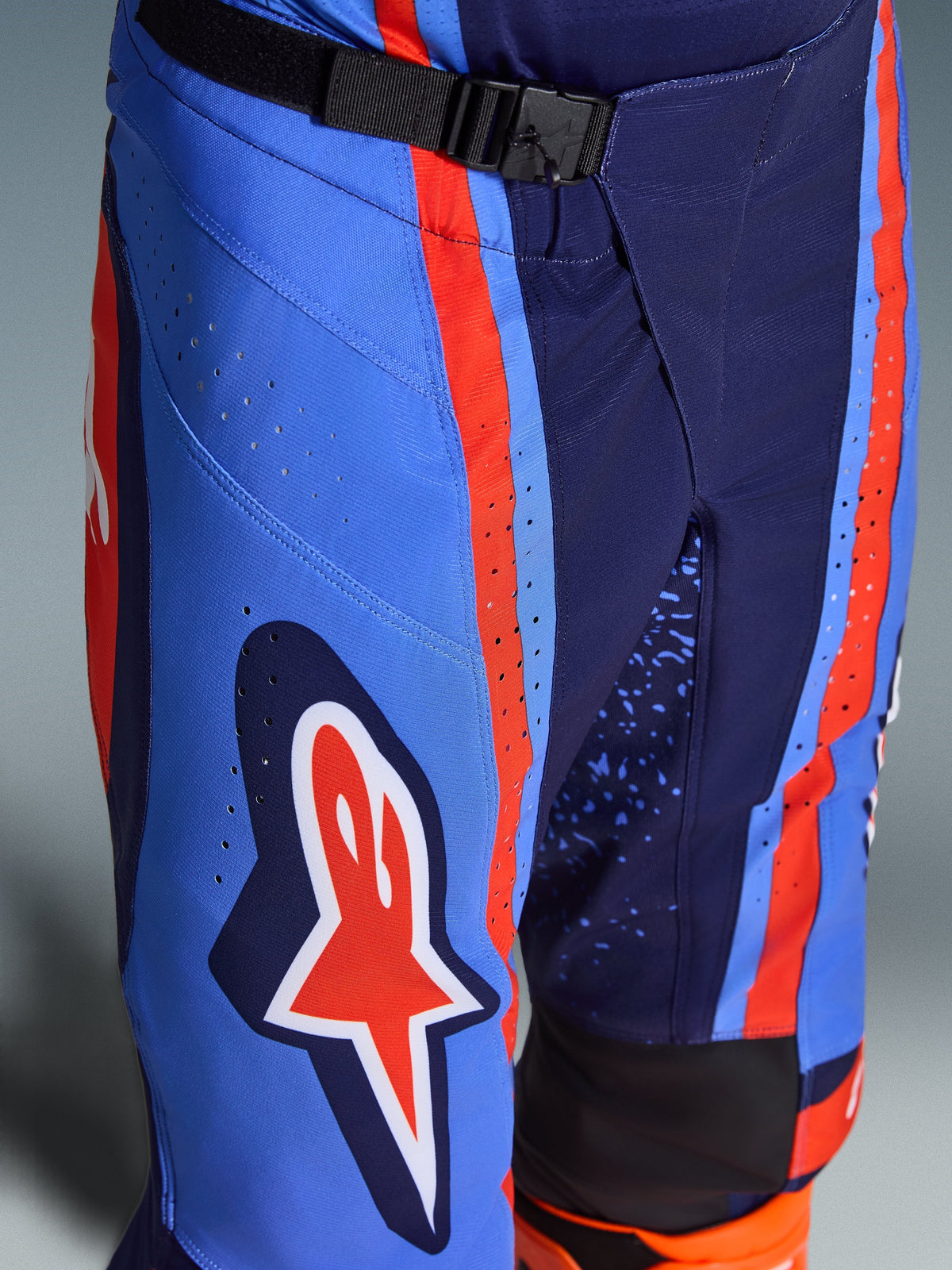Alpinestars 2026 Techstar Nomur Pants, Motocross Pants in Dark Navy Orange and UCLA Blue, close-up showing the SX2 magnetic buckle waist closure, laser-cut perforations on the thigh, and iconic star logo, durable off-road racing gear.