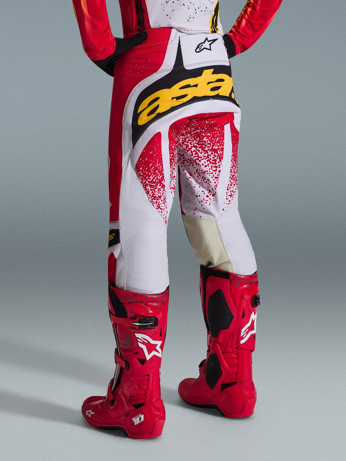 Vista trasera de un piloto de motocross con pantalones Alpinestars 2026 Techstar Nomur en rojo y blanco, con un llamativo logo amarillo en el asiento, combinados con botas de motocross Tech 10 en color rojo brillante, toma de estudio sobre fondo gris.