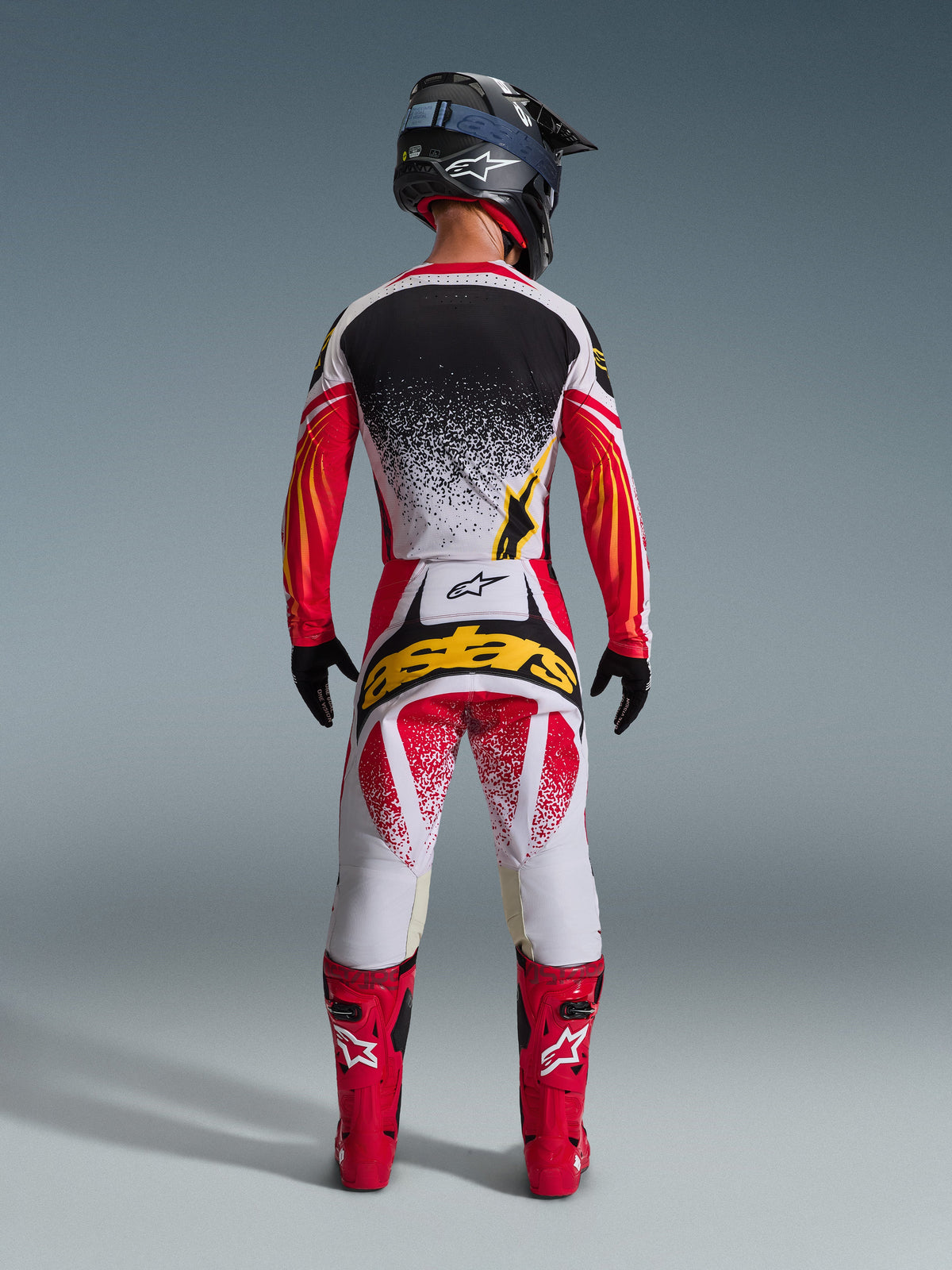 Alpinestars 2026 Techstar Nomur Gear Set, full motocross racing outfit shown from the back, white jersey with black splatter effect and red-orange graphic sleeves, white and red pants with yellow 'astars' logo, red racing boots, black carbon-texture helmet with blue goggles, high-performance off-road gear.