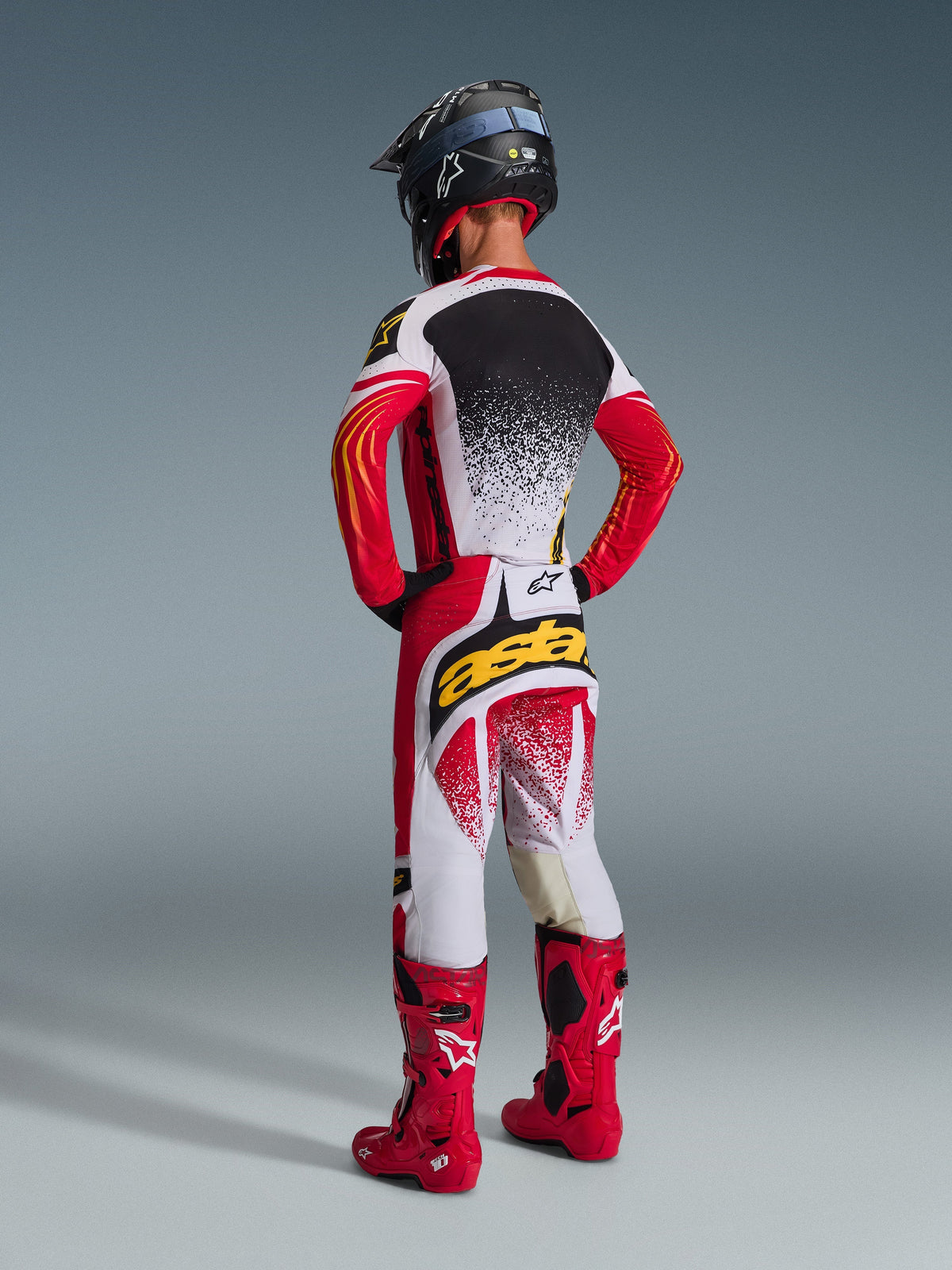 Piloto de motocross visto desde atrás con equipamiento de competición completo Alpinestars, pantalones Techstar Nomur 2026 en rojo, blanco y negro, jersey a juego con diseño moteado, botas rojo brillante, casco negro, logotipo 'astars' destacado en el asiento y conjunto coordinado de carreras todoterreno.