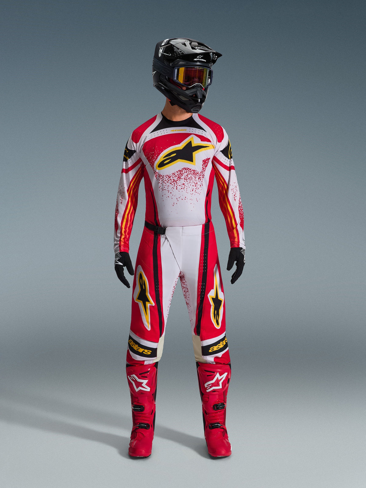 Piloto de motocross con pantalones Alpinestars Techstar Nomur 2026, equipamiento de carreras todoterreno en rojo, blanco y negro, jersey a juego con logo de estrella amarilla, casco negro con gafas, botas rojas y guantes negros, conjunto coordinado para motocross