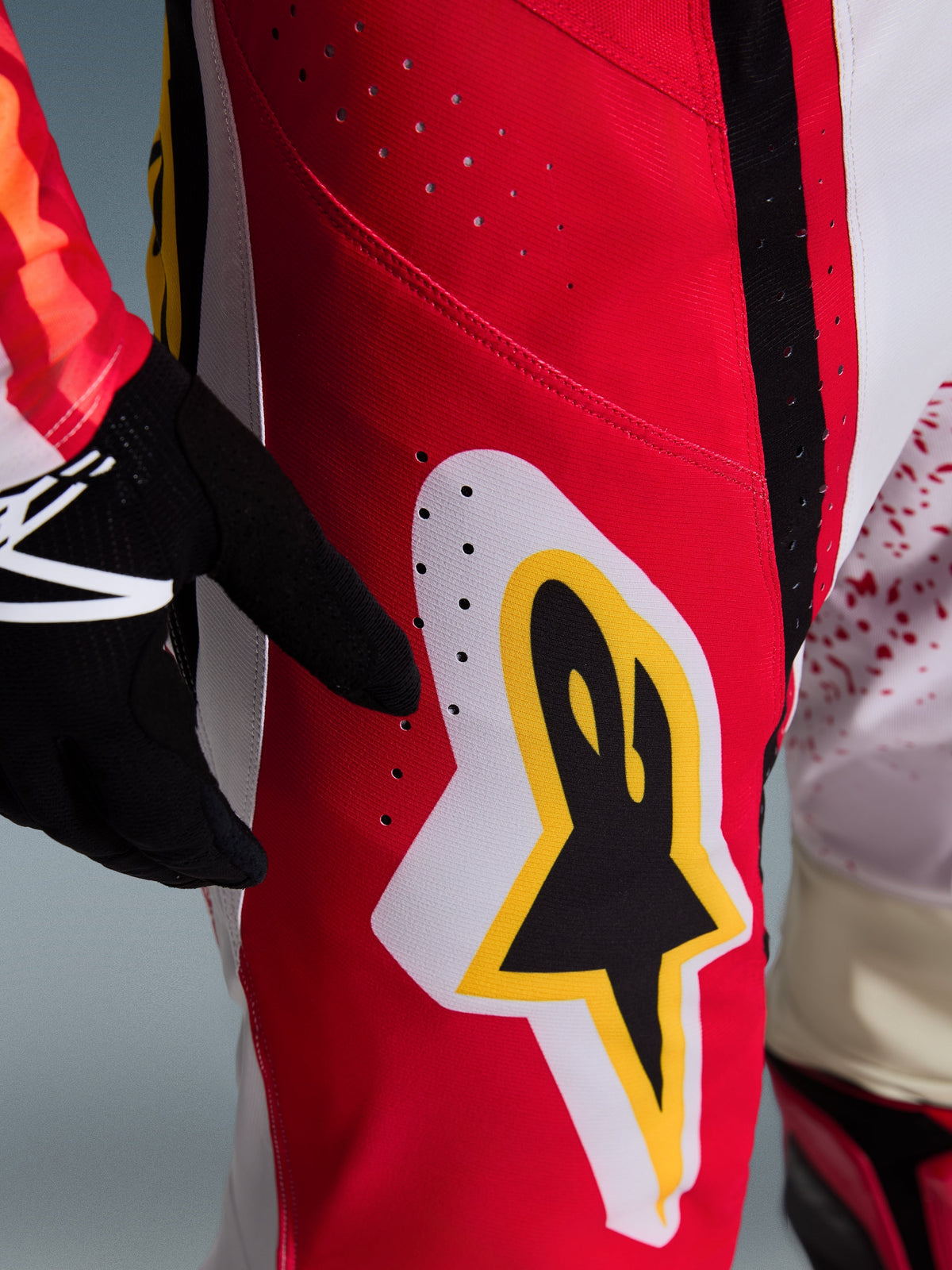Alpinestars Techstar Nomur 2026, pantalones de motocross, rojos con detalles blancos y amarillos, primer plano del área del muslo con orificios de ventilación cortados con láser y un gran logotipo de estrella, combinados con guantes negros y jersey colorido para rendimiento todoterreno