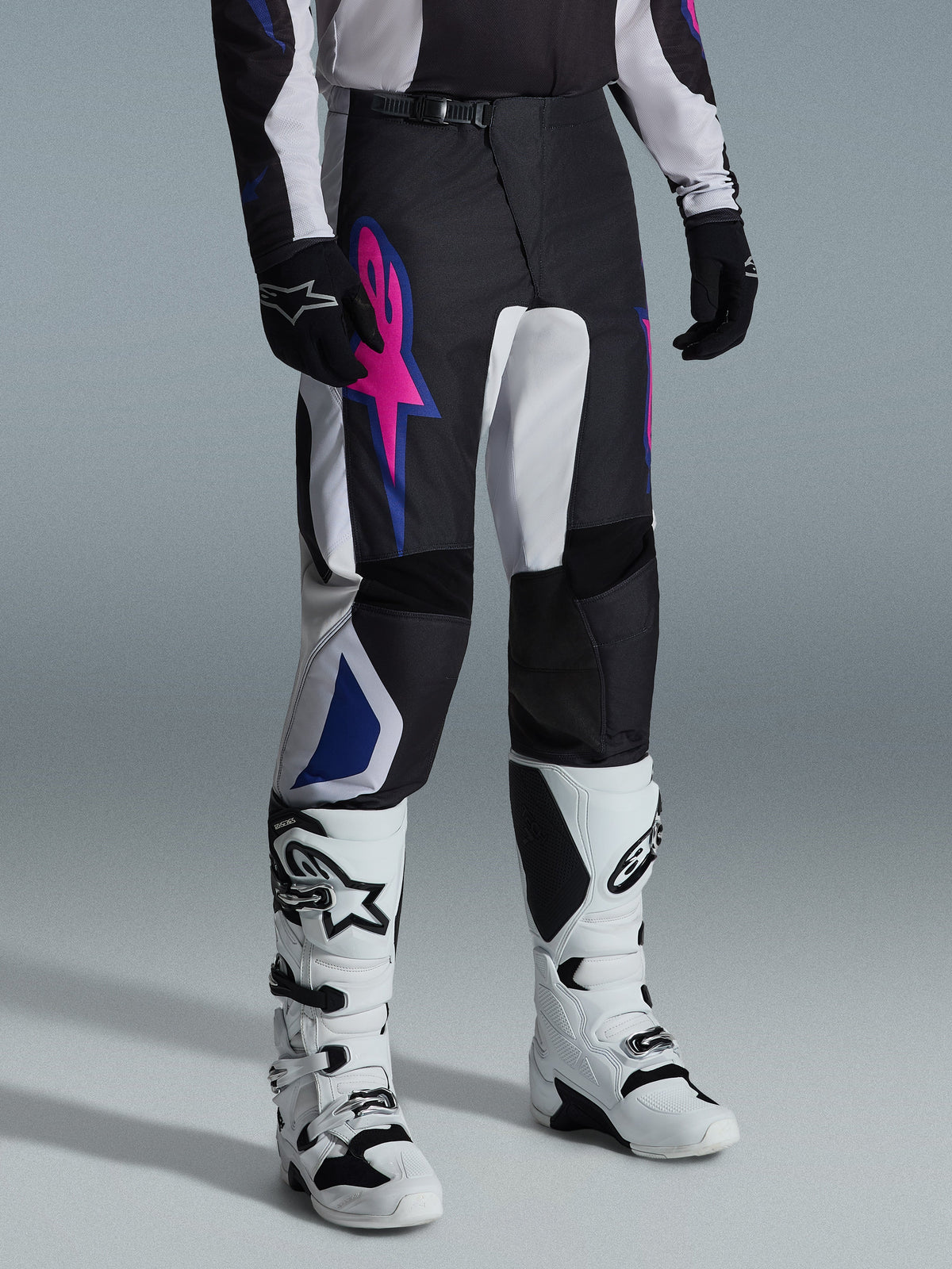 Alpinestars 2026 Fluid Grid Pants, pantalones de motocross, negro, gris claro y púrpura, con un llamativo logotipo de estrella rosa y detalles geométricos azules, usados con botas de motocross blancas y guantes negros, equipamiento ergonómico para conducción todoterreno