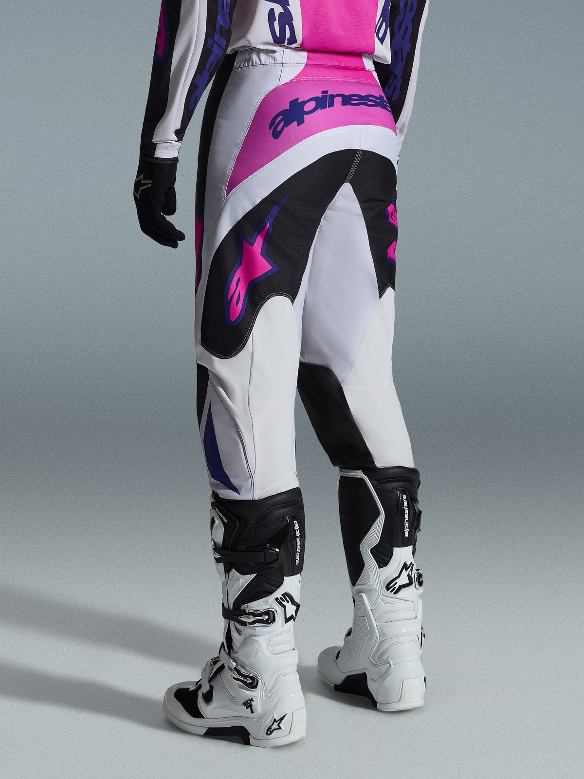 Alpinestars 2026 Fluid Grid Pants, pantalones de motocross, negro, gris claro y púrpura, vista trasera del piloto usando los pantalones con botas blancas y negras, con logotipos de Alpinestars en rosa y púrpura, ajuste atlético para carreras off-road