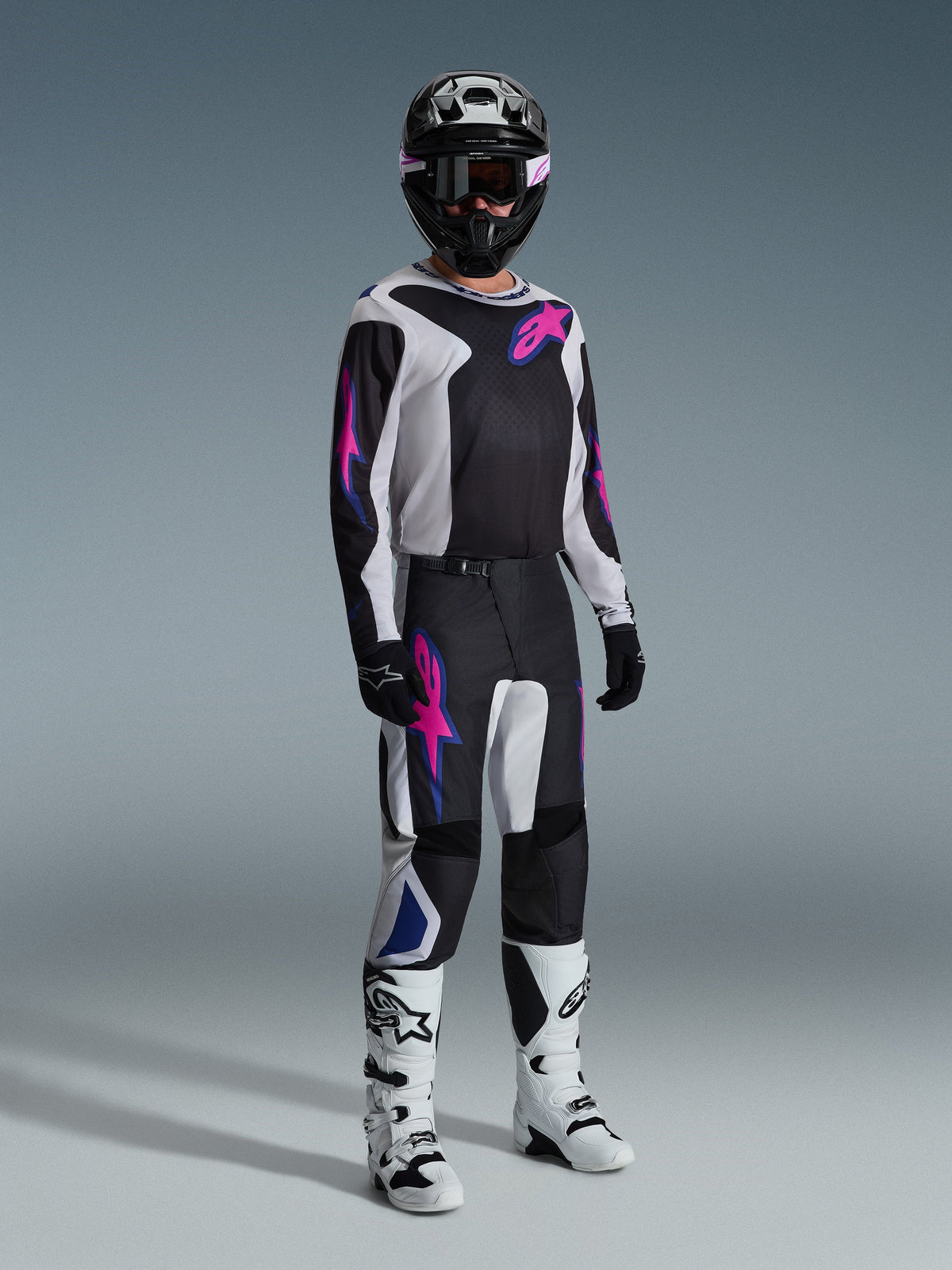 Piloto de motocross con equipamiento de carreras completo, pantalones Alpinestars 2026 Fluid Grid en negro, gris claro y púrpura, jersey a juego con gráficos rosa y azul, casco y gafas negras, botas blancas y negras, y guantes negros, conjunto coordinado para carreras todoterreno