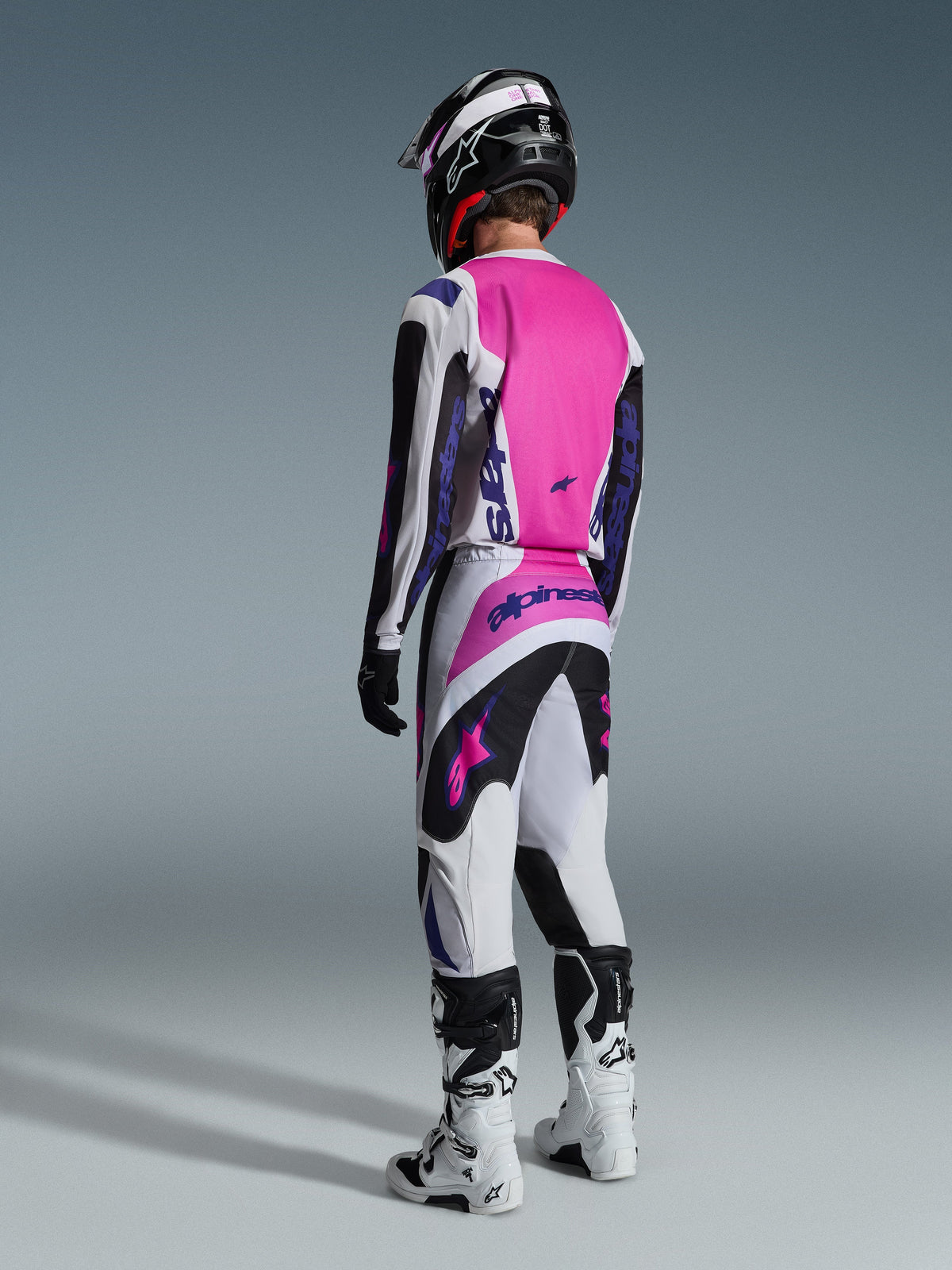 Alpinestars 2026 Fluid Grid Pants, pantalones de motocross, negro, gris claro y morado, vista trasera de un piloto con jersey a juego y botas blancas, con logotipos morados llamativos y paneles rosa vibrante, diseñados para el rendimiento en motocross todoterreno.