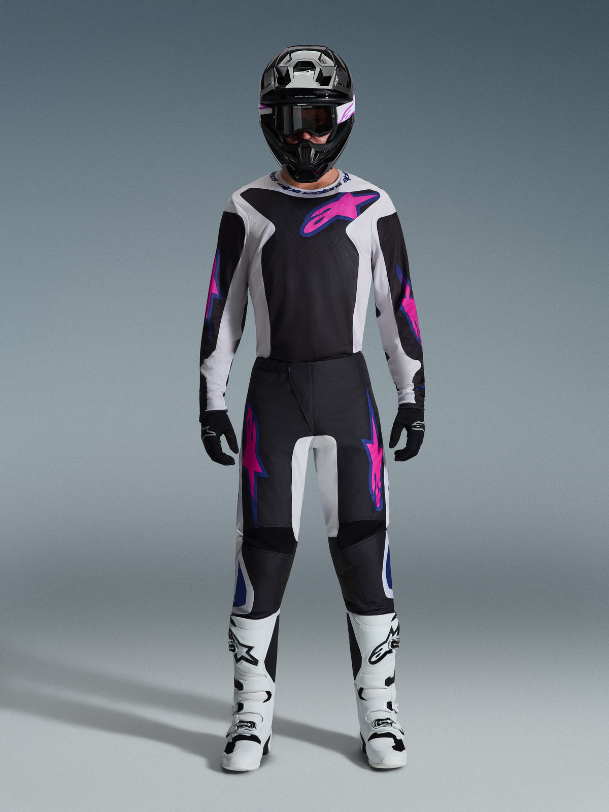 Piloto de motocross con el conjunto Alpinestars Fluid Grid 2026, jersey negro y gris claro con logotipos en rosa brillante, pantalones negros y blancos con gráficos en rosa y azul, casco negro con gafas rosas y botas blancas y negras, equipo completo de competición off-road