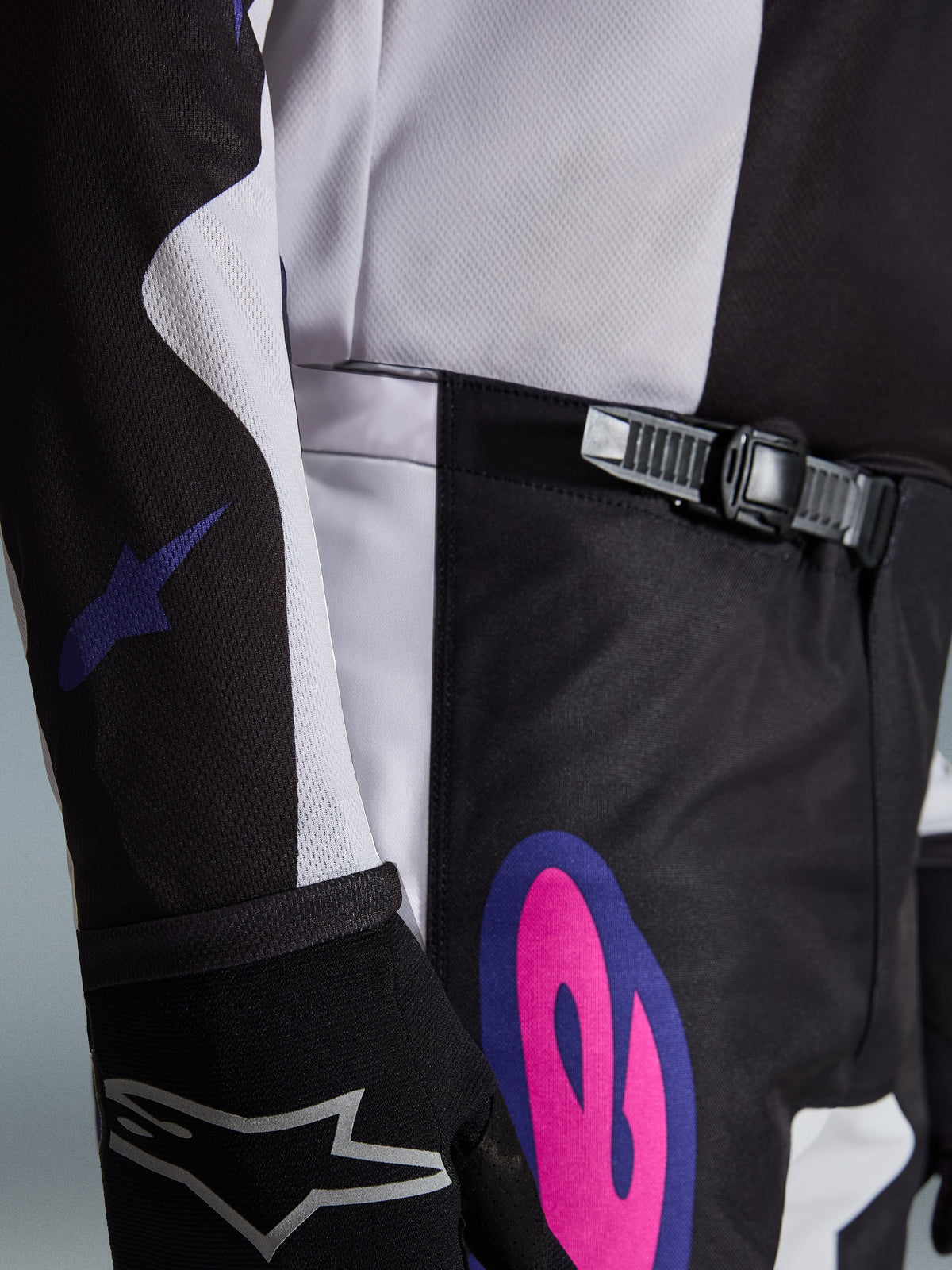 Alpinestars 2026 Fluid Grid Pants, pantalones de motocross, negro, gris claro y morado, primer plano del sistema de cierre de trinquete en la cintura y un gráfico llamativo en rosa y morado en la cadera, usado con un jersey a juego y un guante negro.