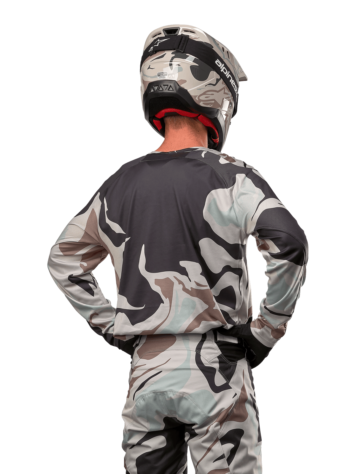 Piloto de motocross visto desde atrás con el Jersey Alpinestars Racer Tactical 2024 en Iron Camo Dust Gray, pantalones y casco de camuflaje a juego, manos en las caderas, equipamiento de carreras todoterreno coordinado.