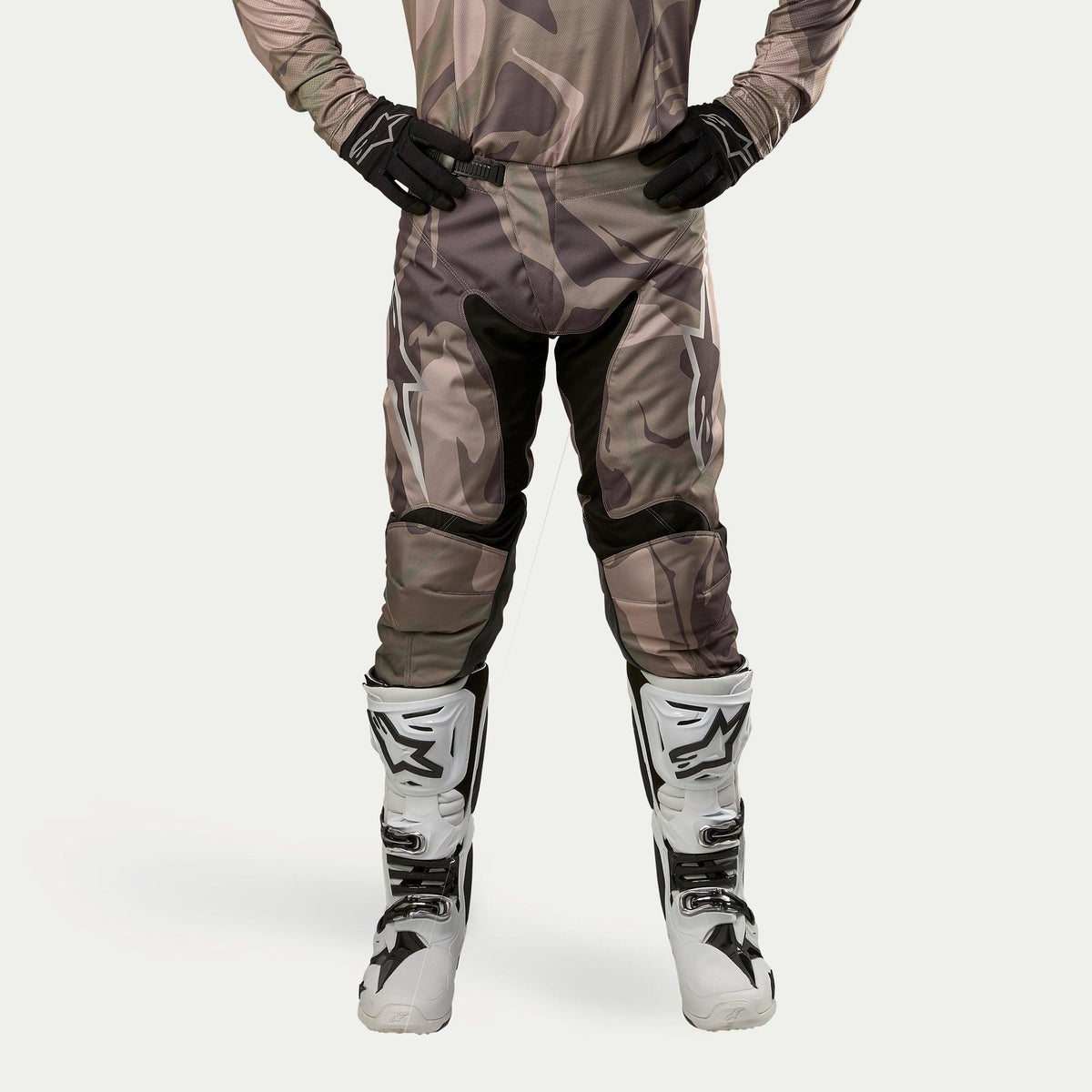 Alpinestars Racer Tactical Pants 2024, pantalones de motocross, verde camuflaje militar marrón, con grandes logotipos Alpinestars blancos en los muslos y paneles interiores de las piernas negros, combinados con botas de motocross blancas y negras, guantes negros y jersey de camuflaje a juego, vista frontal.
