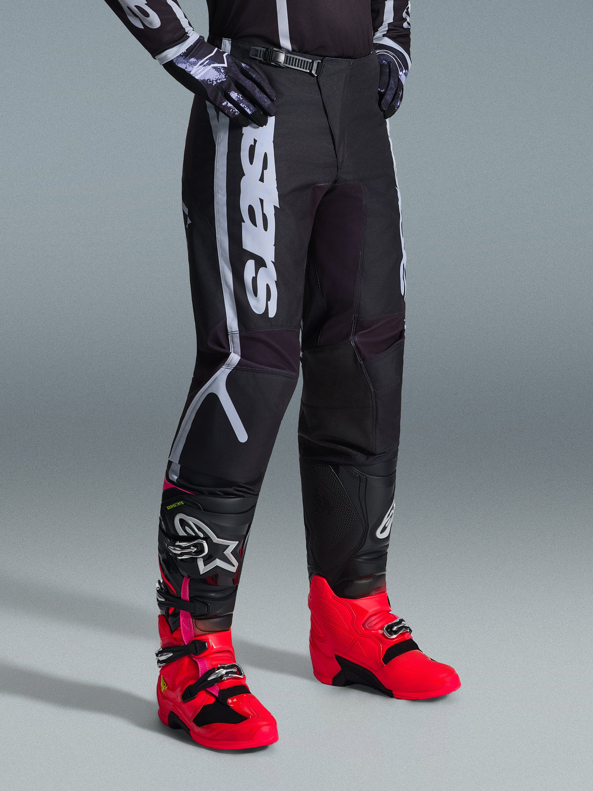 Pantalones Alpinestars Fluid Apex 2026, pantalones de motocross, negros y grises, usados con botas de color rosa vibrante y rojo y jersey a juego en blanco y negro, con el logotipo vertical de Alpinestars en el muslo, paneles reforzados y cierre de trinquete en la cintura para un óptimo rendimiento off-road.