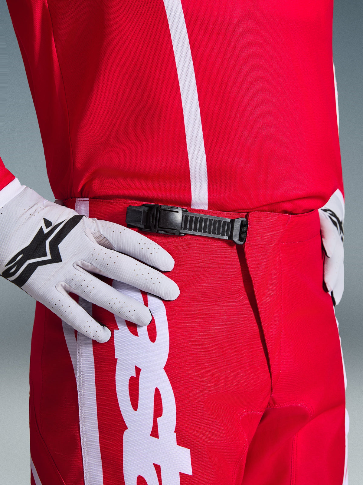 Pantalones Alpinestars Fluid Apex 2026, pantalones de motocross, rojos y blancos, primer plano del sistema de cierre de trinquete negro en la cintura, usados con guantes Alpinestars blancos y jersey rojo a juego, equipamiento de alto rendimiento duradero para conducción off-road