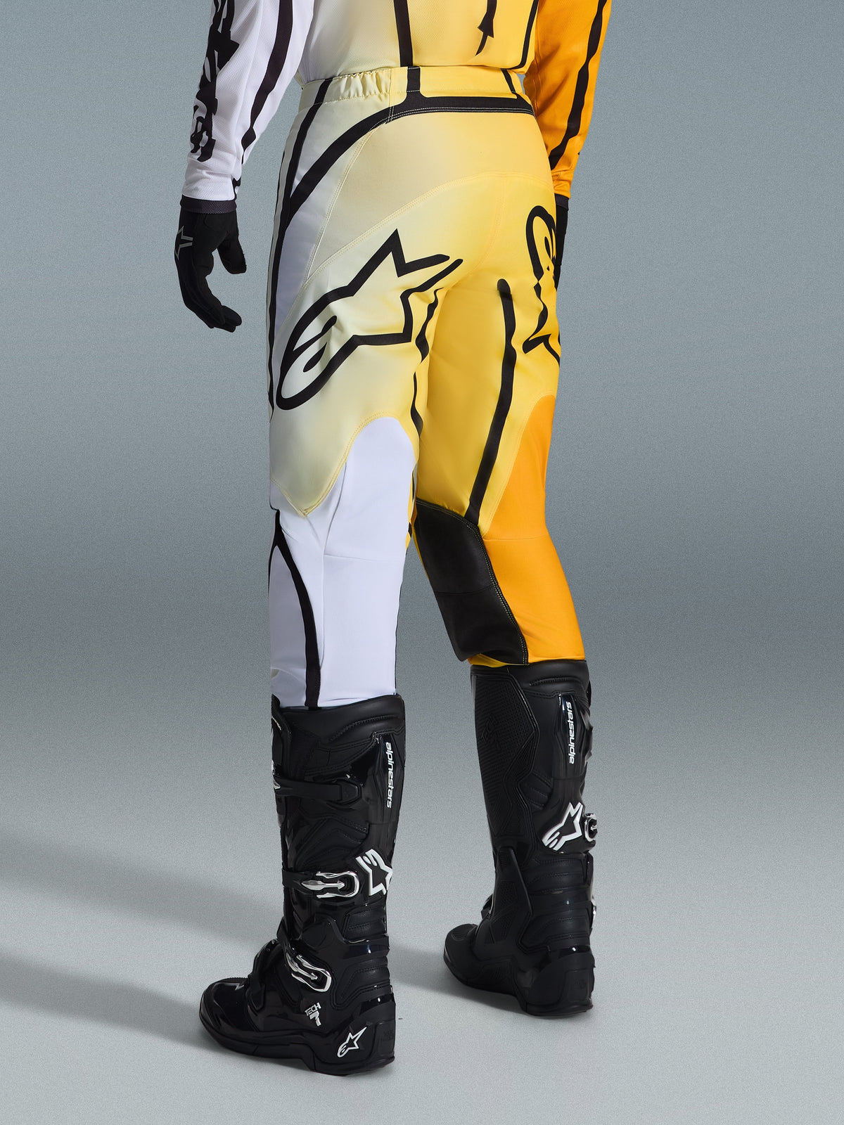 Alpinestars 2026 Fluid Apex Pants, pantalones de motocross con degradado en amarillo y negro y detalles blancos y naranjas, vista desde atrás con jersey a juego y botas Tech 7 negras, con un llamativo logotipo negro de Alpinestars en la parte trasera, equipamiento de alto rendimiento para conducción off-road
