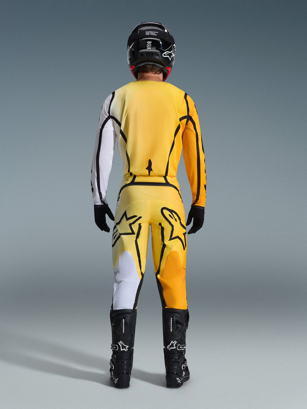 Alpinestars 2026 Fluid Apex Pants, pantalones de motocross, amarillos y negros, vista trasera de un piloto con equipamiento completo de motocross, jersey a juego con degradados blancos y naranjas, casco negro, botas negras y guantes negros, conjunto coordinado para todoterreno.
