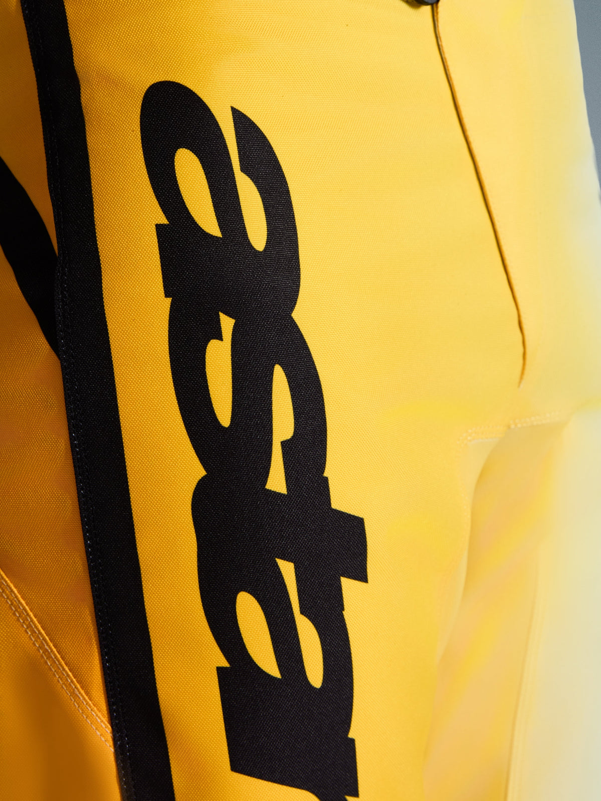 Primer plano de Alpinestars 2026 Fluid Apex Pants, pantalones de motocross, amarillo y negro, con un gran logotipo negro vertical, material duradero texturizado y detalles ergonómicos para un rendimiento todoterreno