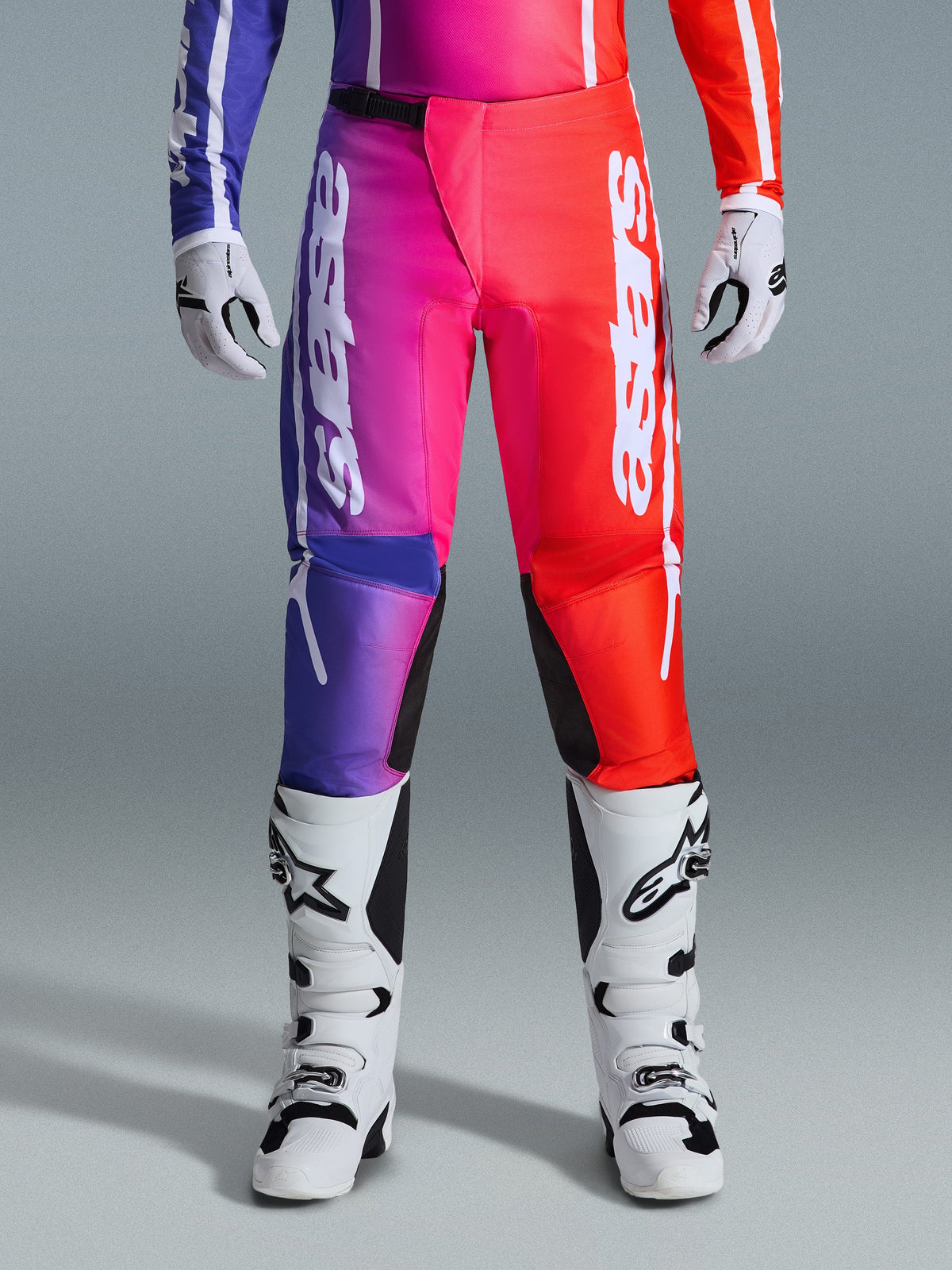Pantalones Alpinestars Fluid Apex 2026, pantalones de motocross, diseño con degradado multicolor en púrpura, rosa y rojo, logotipo grande de Alpinestars en blanco en las piernas, combinados con botas y guantes de motocross blancos y negros, equipamiento profesional para carreras todoterreno