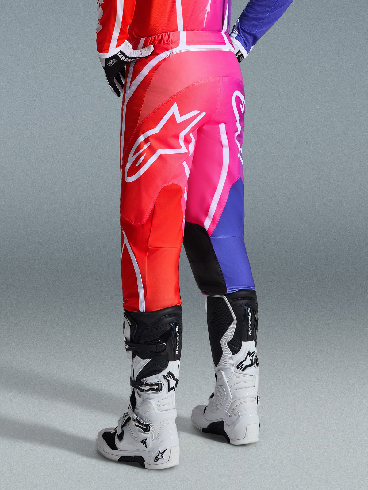 Pantalones Alpinestars 2026 Fluid Apex, pantalones de motocross, multicolor rojo, rosa y púrpura, vista trasera de piloto con botas de motocross blancas y negras, con logo grande de Alpinestars en blanco y diseño de degradado vibrante