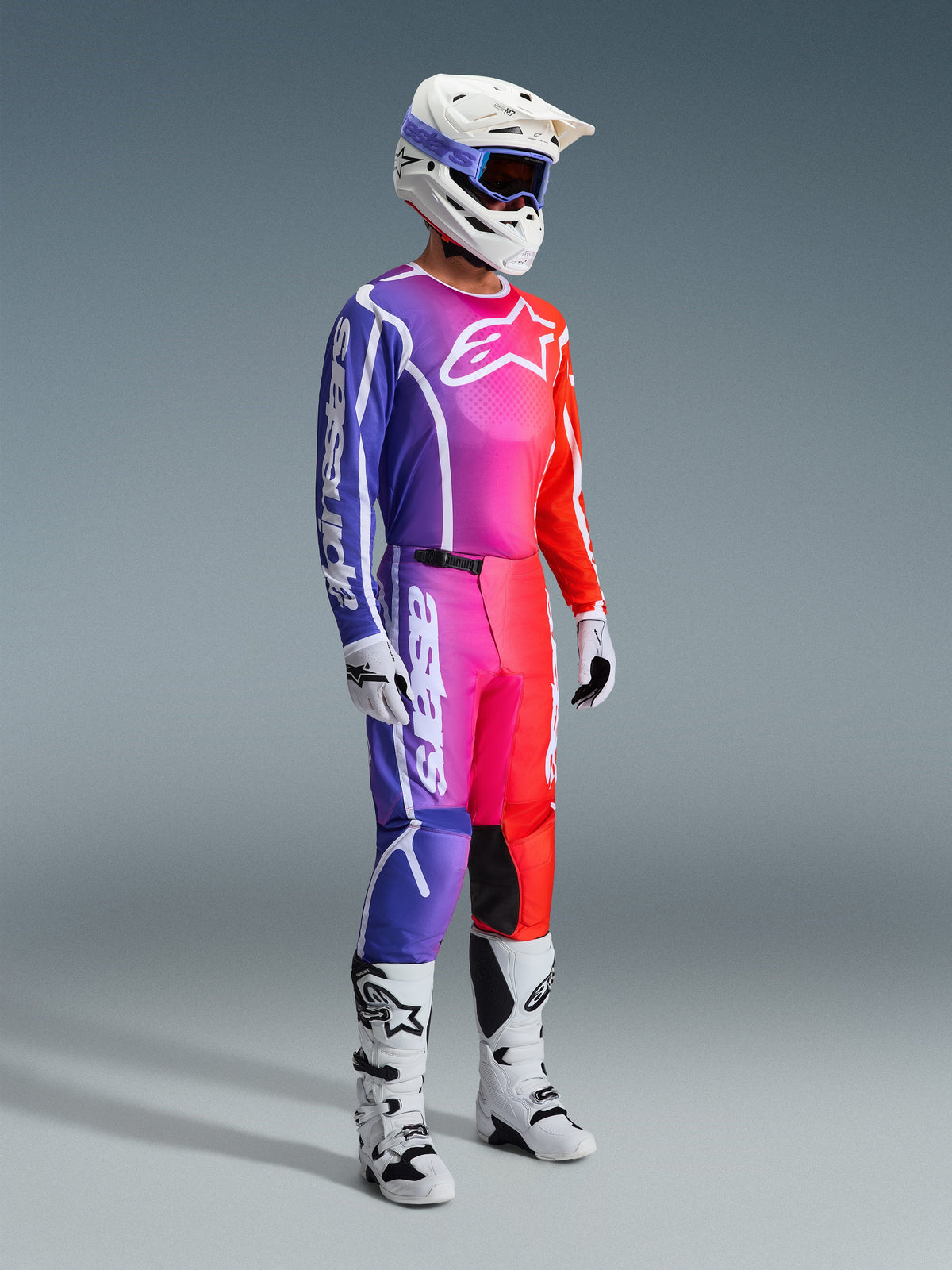 Pantalones Alpinestars Fluid Apex 2026, pantalones de motocross, degradado multicolor de púrpura, rosa y naranja, usados con jersey a juego, casco blanco y botas blancas, con logotipos blancos llamativos y diseño ergonómico para carreras todoterreno