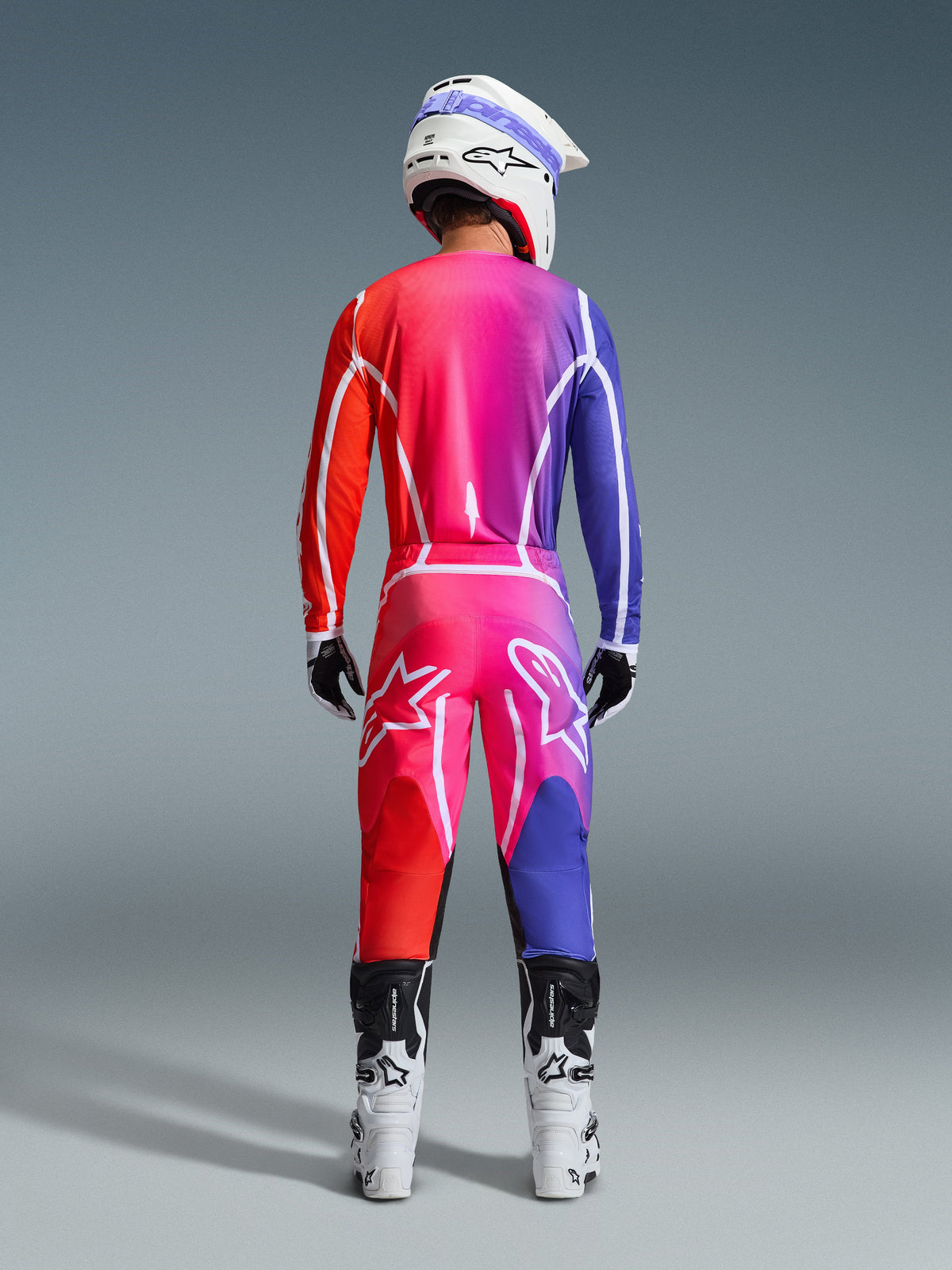 Vista posterior de un piloto de motocross con pantalones Alpinestars 2026 Fluid Apex y jersey a juego, con un vibrante degradado en rojo, rosa y morado, combinado con un casco blanco y botas blancas, conjunto de equipamiento coordinado para carreras off-road