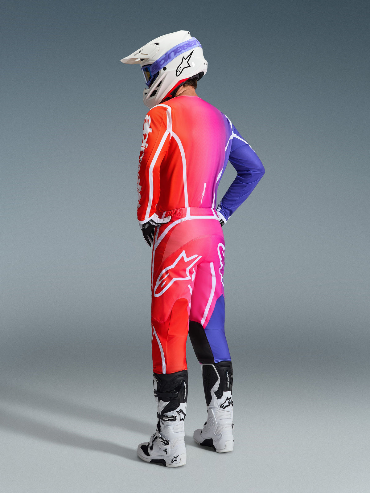 Piloto de motocross con equipamiento Alpinestars 2026 Fluid Apex, jersey y pantalones multicolores con un degradado en rojo, rosa y morado y logotipos blancos, casco blanco y botas blancas, conjunto coordinado para carreras todoterreno
