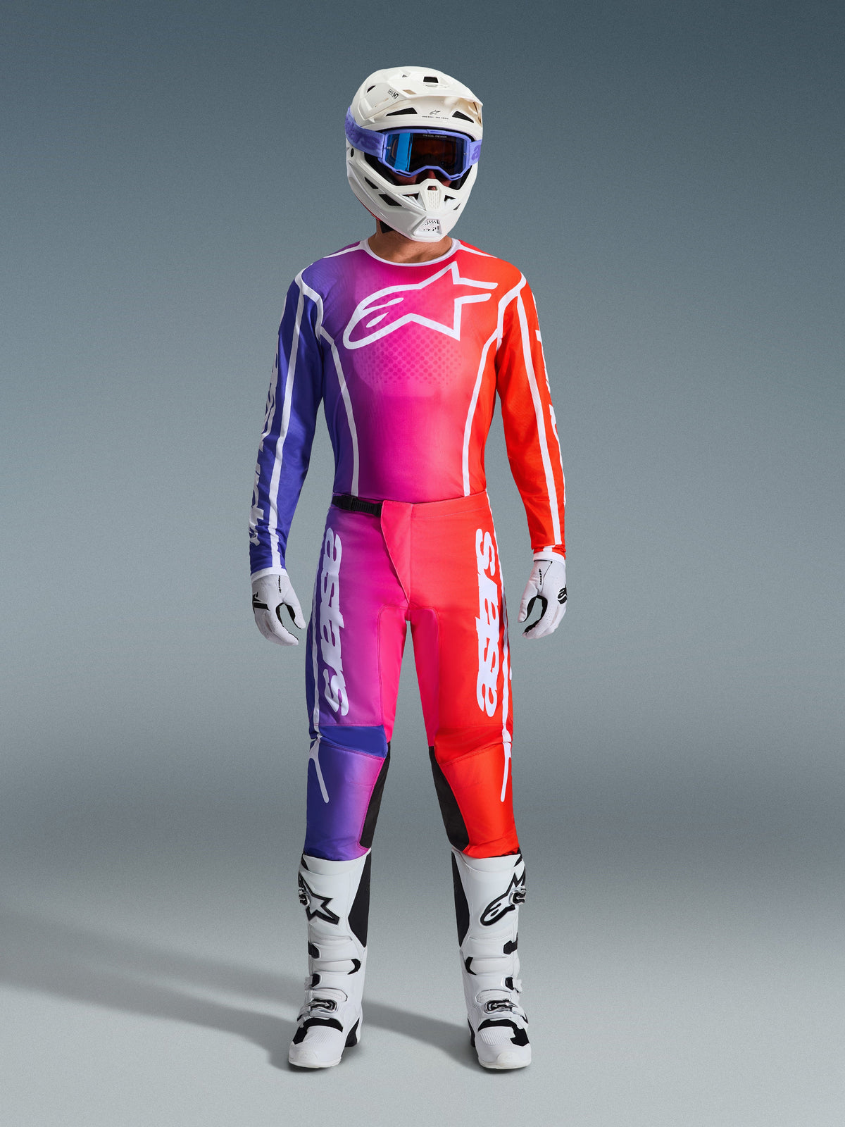Piloto de motocross con pantalones Fluid Apex 2026 y jersey a juego, degradado de púrpura y naranja, casco blanco con gafas, botas y guantes blancos, equipamiento de carreras Alpinestars coordinado para rendimiento off-road.