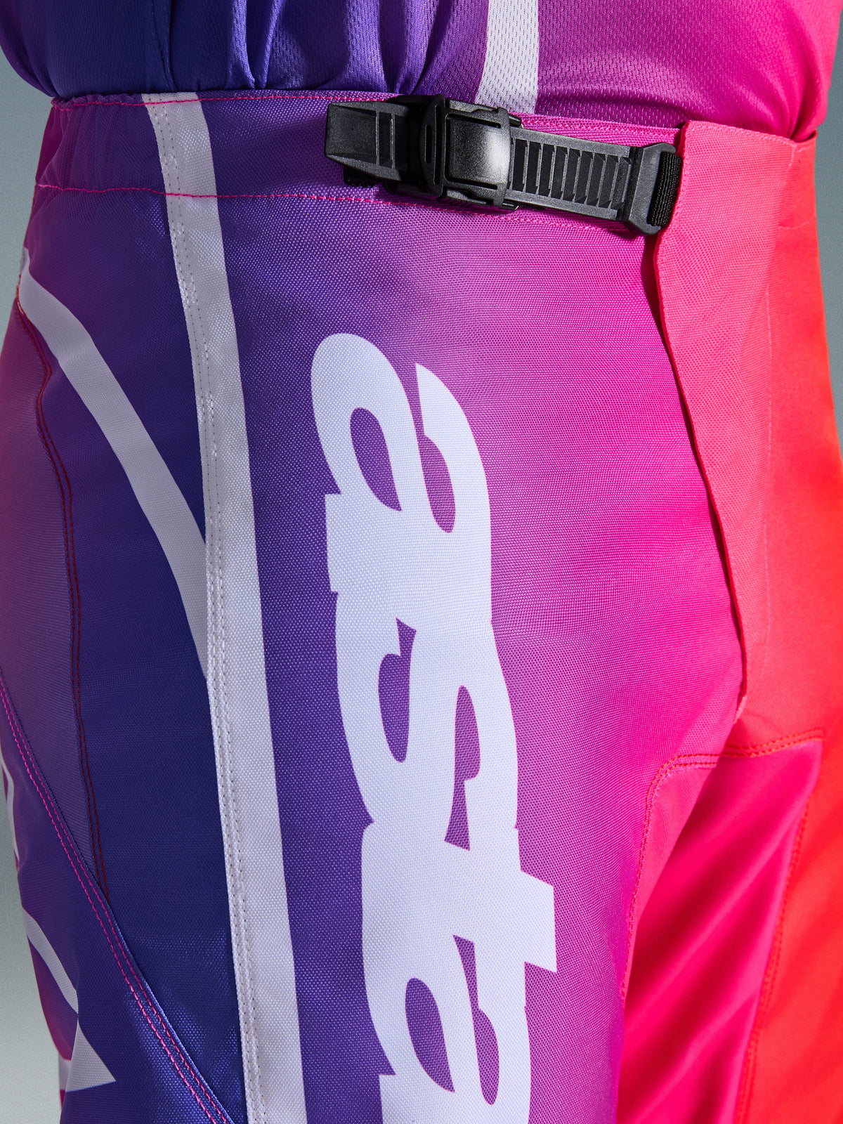 Alpinestars 2026 Fluid Apex Pants, pantalones de motocross, multicolor púrpura, rosa y rojo, primer plano de la zona de la cintura y la cadera con sistema de cierre de trinquete negro y gran logotipo vertical de Alpinestars en blanco, diseño con degradado vibrante y textura de polifabric duradera