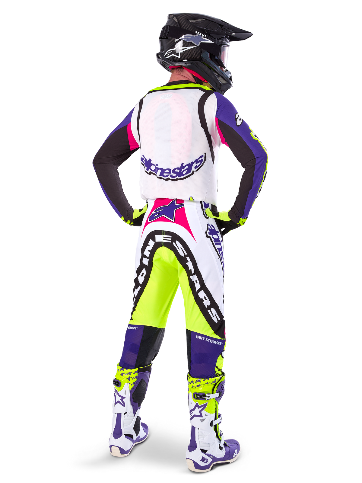 Piloto de motocross con pantalones Alpinestars Supertech Dirt Studios® de Edición Limitada en blanco, púrpura y amarillo flúor, visto desde atrás con jersey y botas a juego y casco de carbono negro, equipamiento profesional de carreras off-road con gráficos llamativos y detalles en neón.