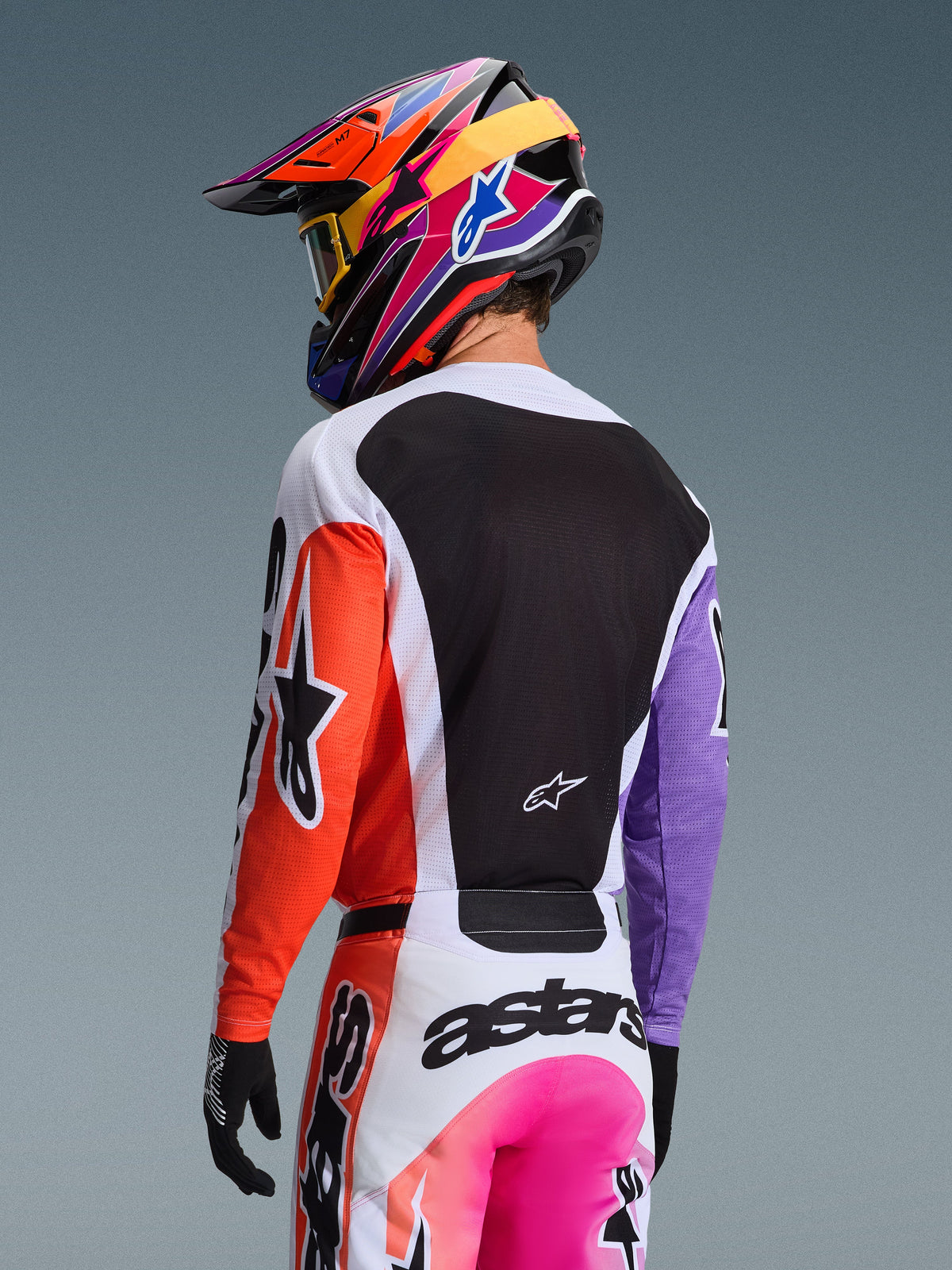 Piloto de motocross visto desde atrás con el Jersey Alpinestars Racer Air Portl 2026 Multicolor, con manga derecha naranja, manga izquierda morada y un amplio panel trasero de malla negra para ventilación, combinado con pantalones de motocross a juego y un vibrante casco multicolor, equipamiento profesional para carreras off-road.