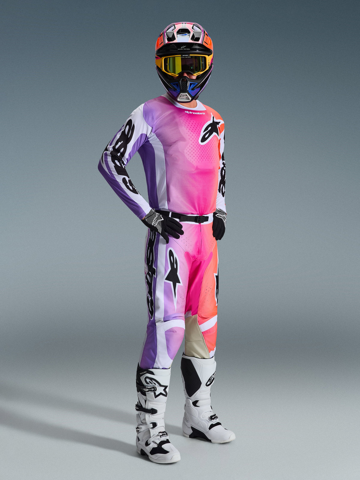 Piloto de motocross con el Jersey Alpinestars 2026 Racer Air Portl y pantalones a juego, degradado multicolor vibrante en rosa, púrpura y naranja con detalles blancos, casco colorido coordinado y gafas amarillas, botas blancas y negras, equipamiento completo de carreras para rendimiento off-road