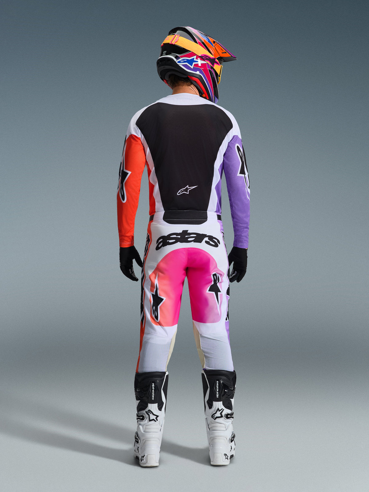 Piloto de motocross con equipamiento completo visto desde atrás, Jersey Alpinestars Racer Air Portl 2026 con paneles blancos, negros, naranjas y morados, pantalones blancos a juego con detalles rosas y marca 'astars', botas blancas y negras, y casco multicolor vibrante, conjunto coordinado para carreras de motocross