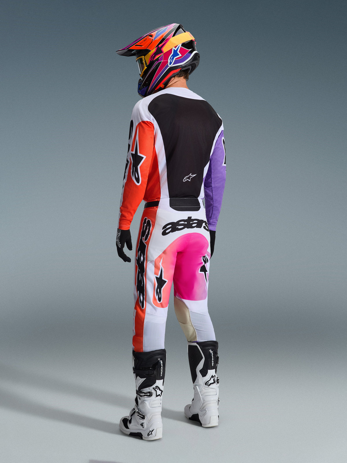 Alpinestars 2026 Racer Air Portl Jersey, jersey de motocross multicolor, vista trasera de un piloto con equipamiento completo de competición, con base blanca, mangas naranjas y moradas y panel trasero de malla negra, combinado con pantalones rosa y naranja a juego, casco colorido y botas blancas, atuendo profesional de competición off-road