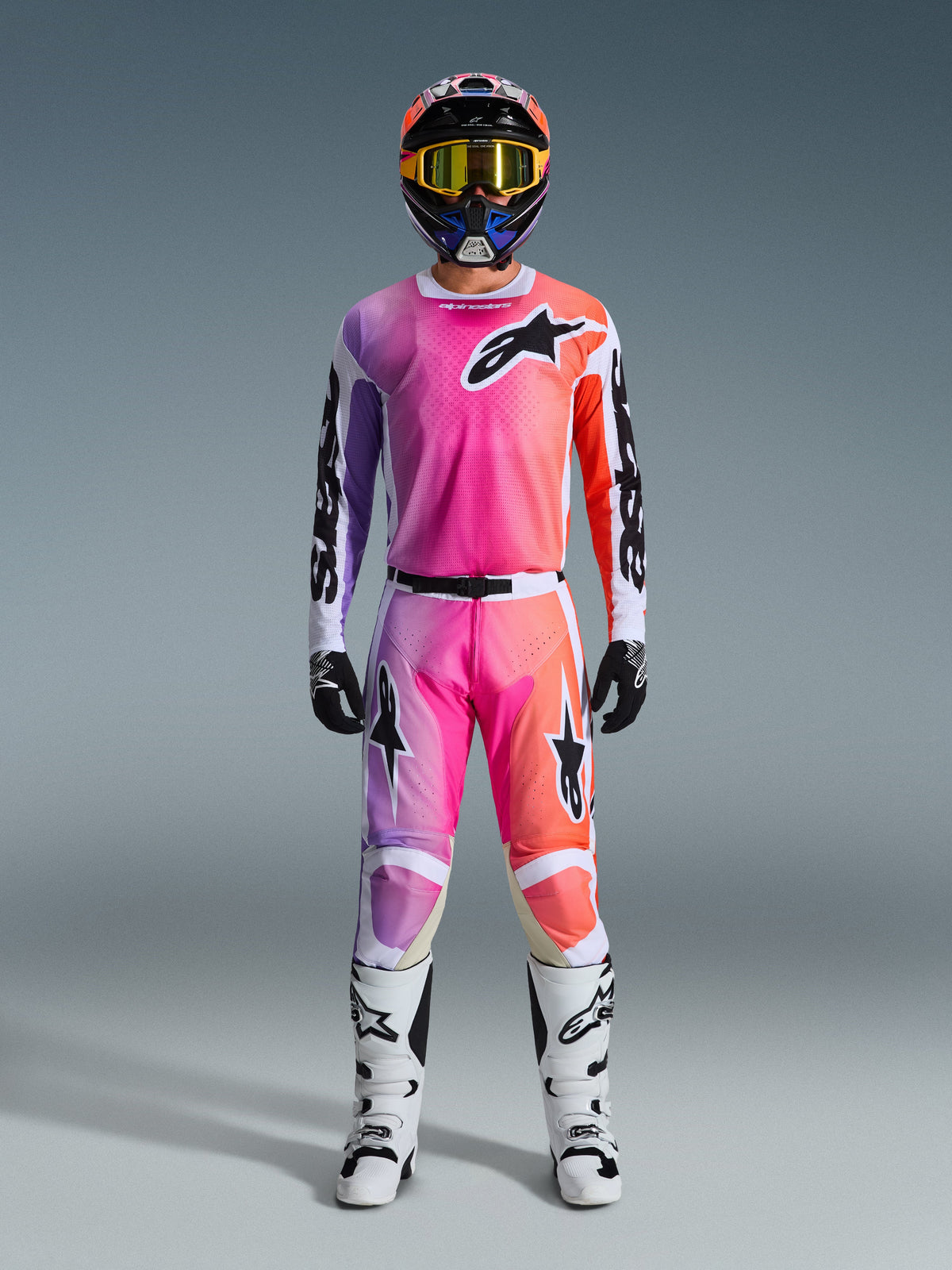 Piloto de motocross con el Jersey Racer Air Portl 2026 y pantalones a juego, con un degradado en rosa, púrpura y naranja con logotipos negros de Alpinestars, botas de competición blancas, guantes negros y casco multicolor, equipamiento profesional de carreras off-road