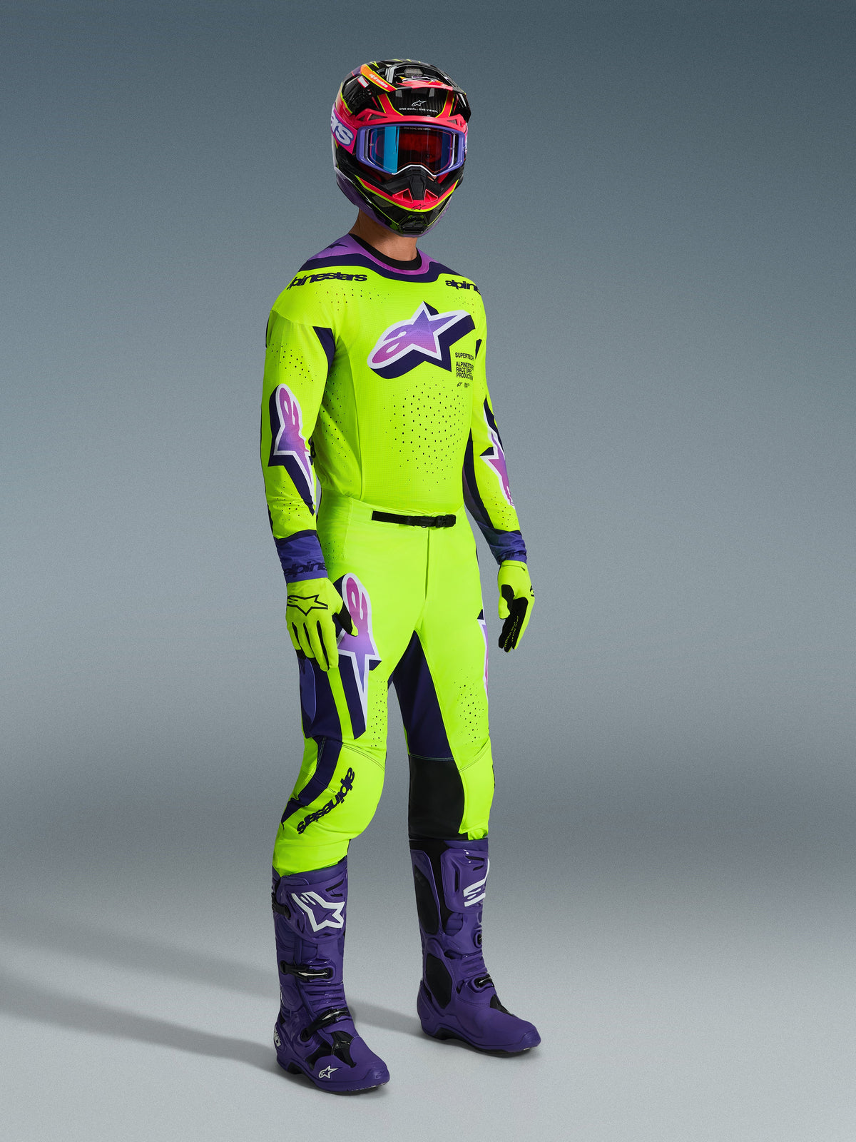 Alpinestars 2026 Supertech Vista Gear Set, piloto profesional de motocross vistiendo un jersey y pantalones en verde lima vibrante con acentos y logotipos morados, guantes verde lima a juego, botas moradas y casco multicolor con gafas azules, equipo coordinado de ajuste atlético para carreras todoterreno