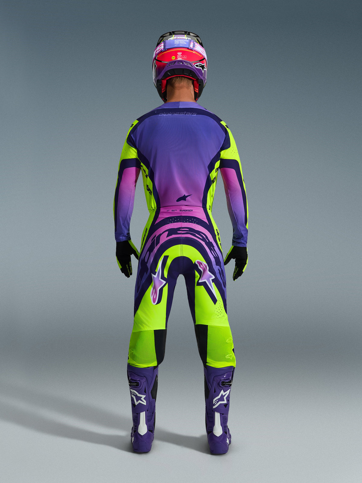 Alpinestars 2026 Supertech Vista Gear Set, vista trasera de un piloto de motocross, traje de carreras con un degradado vibrante en púrpura, rosa y verde lima, con jersey y pantalones a juego con logotipos llamativos, casco coordinado en púrpura y rosa, botas púrpuras y guantes negros.
