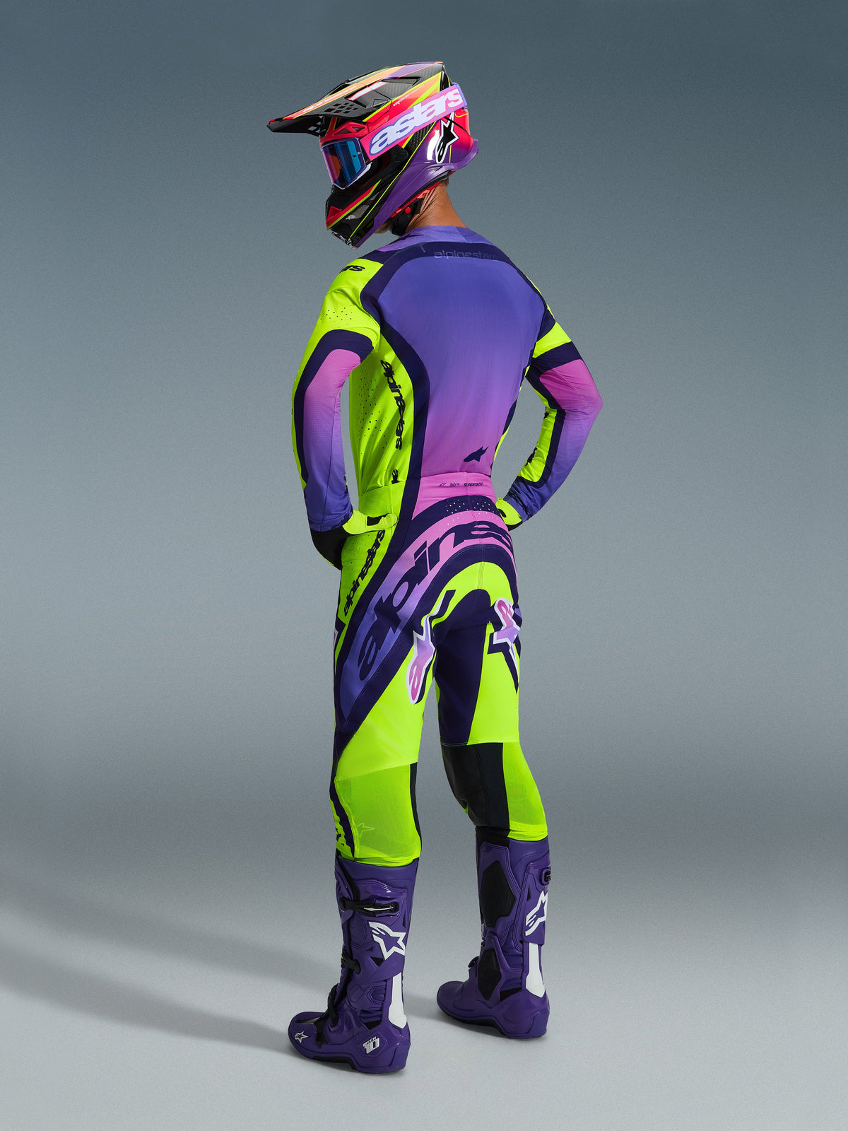 Piloto de motocross desde atrás con el conjunto Alpinestars 2026 Supertech Vista, jersey morado con degradado rosa y detalles en amarillo neón, pantalones a juego en amarillo neón y morado, botas moradas con logotipos blancos, casco y gafas coordinados, ropa de carreras de motocross de nivel profesional