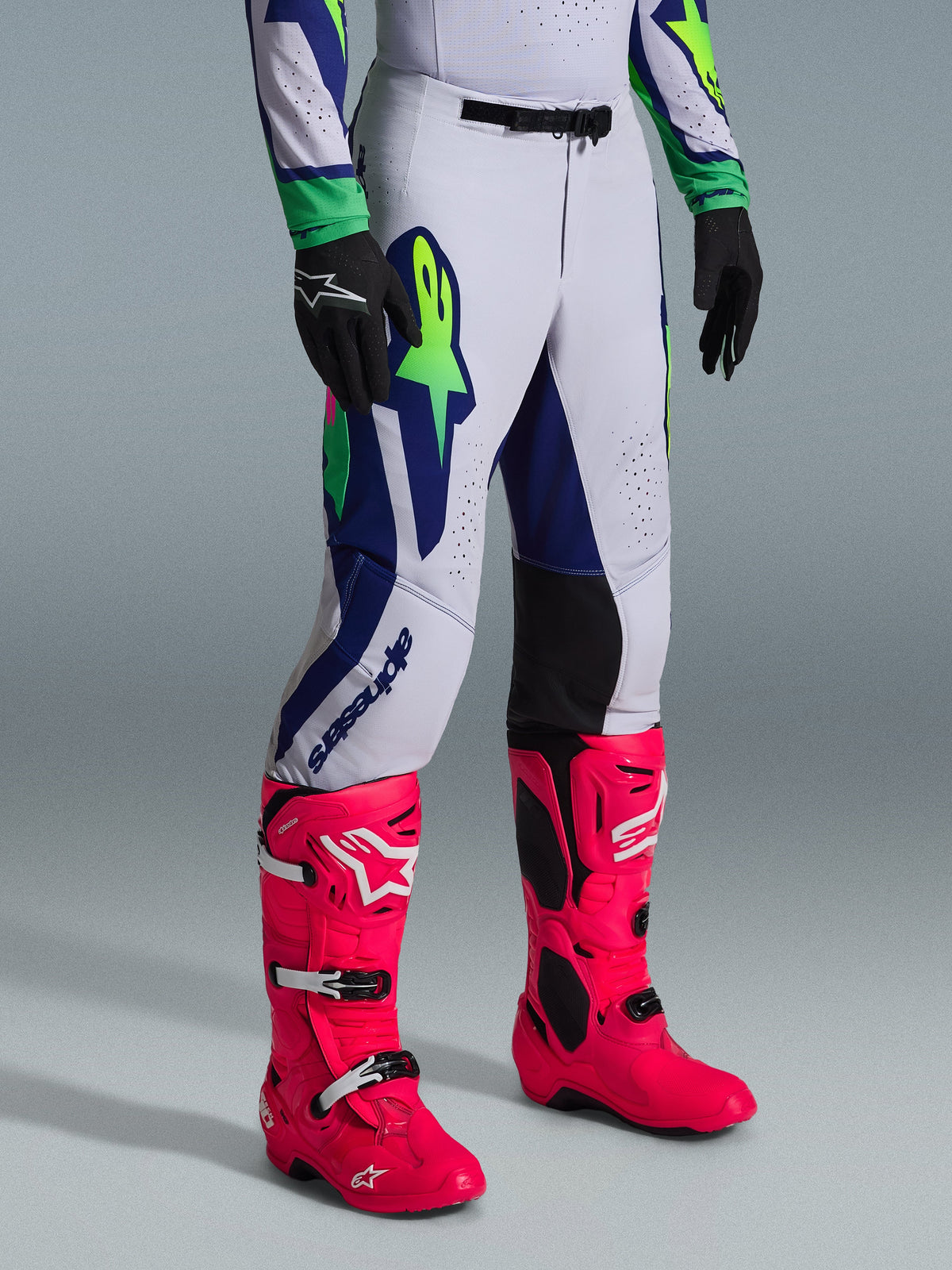 Alpinestars 2026 Supertech Vista Gear Set, pantalones de motocross, gris claro con acentos en azul marino y verde lima, usados con botas rosa brillante y guantes negros, con un ajuste atlético profesional y gráficos llamativos