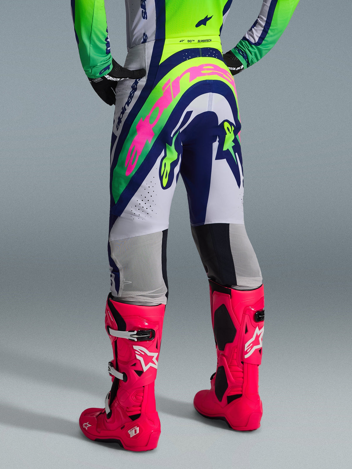 Alpinestars 2026 Supertech Vista Pants, pantalones de motocross, gris claro, amarillo flúor y azul, vista trasera con un gran logotipo rosa de Alpinestars, combinados con jersey de carreras a juego y botas Tech 10 en rosa brillante, equipación profesional para todoterreno