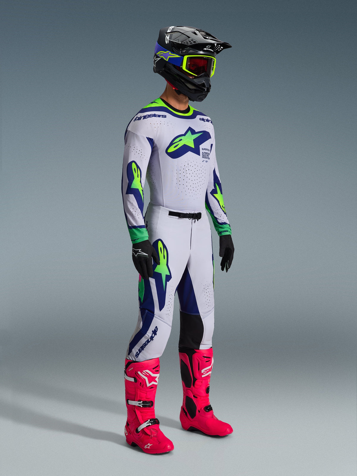 Pantalones Alpinestars 2026 Supertech Vista y jersey a juego, equipamiento de motocross en gris claro con detalles en amarillo flúor y azul, usado por un piloto con casco negro y botas rosa vibrante, con gráficos atléticos y ajuste de competición profesional