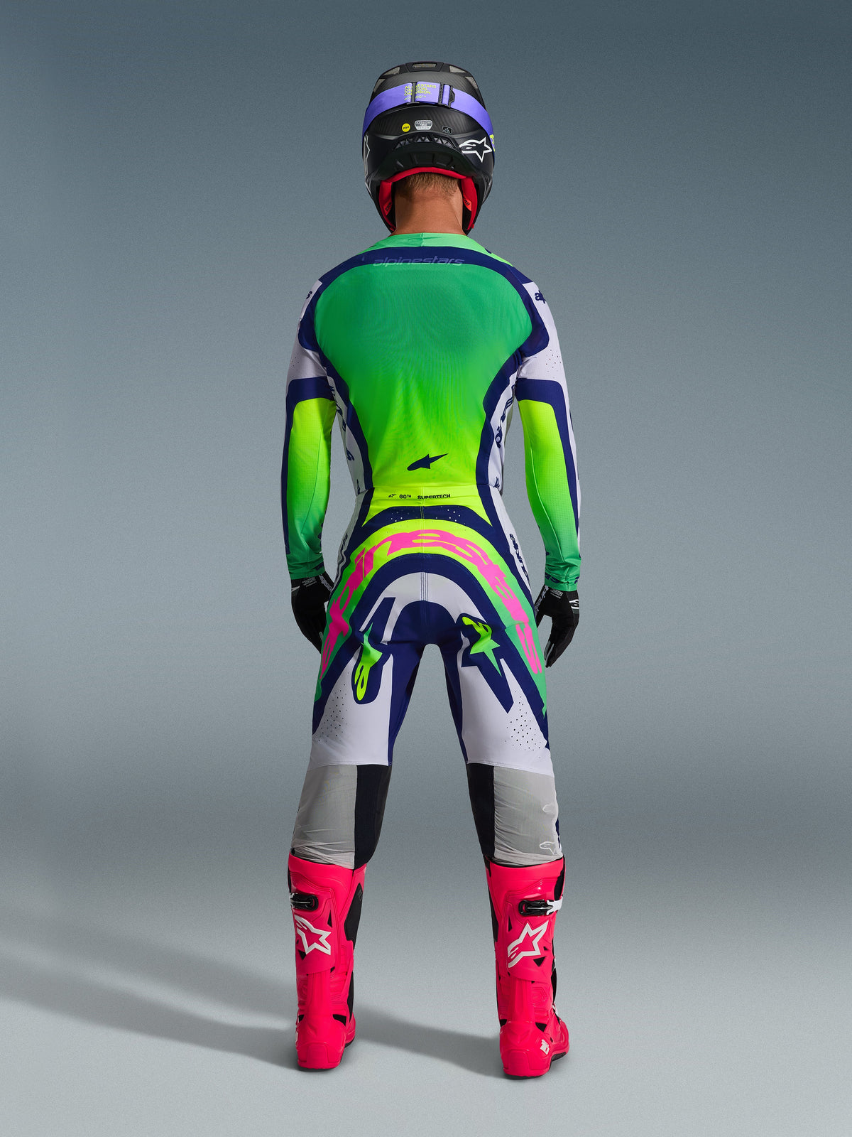 Alpinestars 2026 Supertech Vista Gear Set, vista trasera de conjunto de motocross, jersey con degradado verde y amarillo, pantalones blancos y azules con un gran logo rosa en la parte trasera, botas rosa brillante y casco morado, ropa profesional de carreras todoterreno
