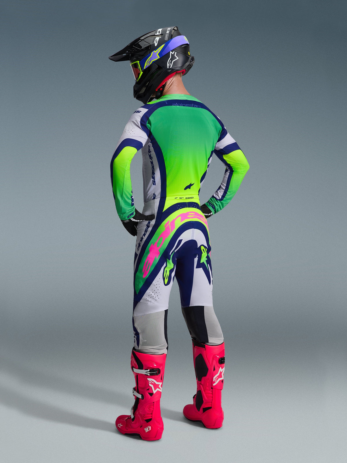 Piloto de motocross visto desde atrás con los pantalones Alpinestars 2026 Supertech Vista, diseño en gris claro, azul y amarillo neón con un logotipo rosa llamativo en el asiento, combinado con un jersey a juego, casco negro y botas rosa brillante para una equipación completa de carreras off-road.