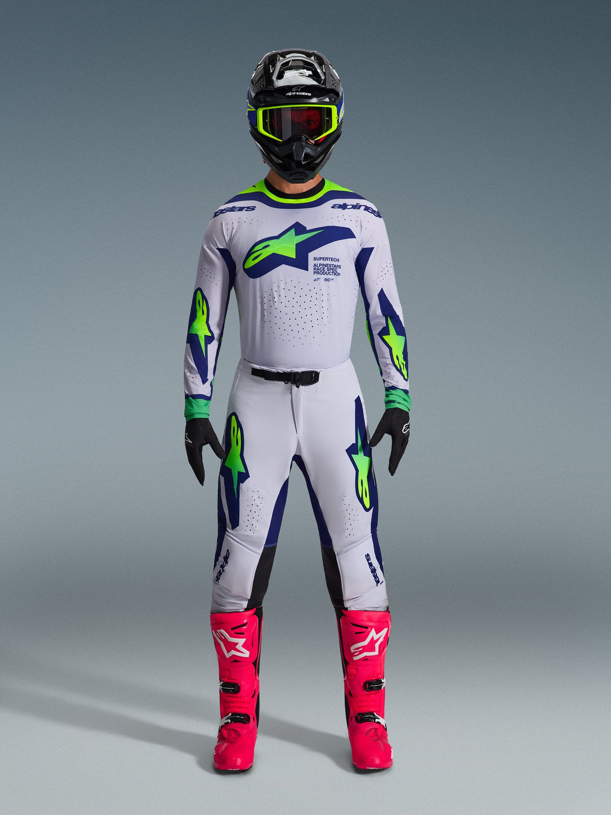 Piloto de motocross con pantalones Alpinestars 2026 Supertech Vista y jersey a juego, color gris claro con acentos en amarillo flúor y azul, ajuste atlético, combinado con casco negro, gafas amarillas y botas rosas vibrantes, conjunto de equipamiento profesional para carreras todoterreno