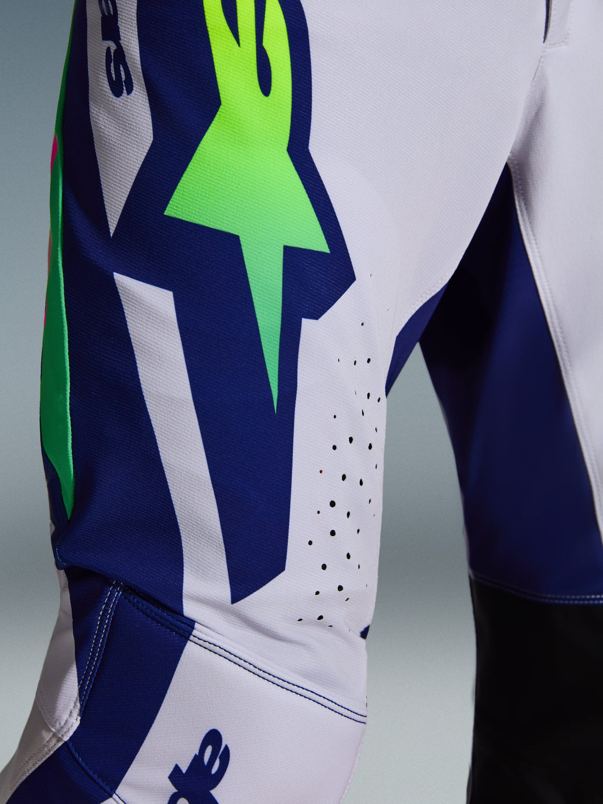 Alpinestars 2026 Supertech Vista Pants, pantalones de motocross, gris claro, amarillo flúor y azul, detalle en primer plano de la zona del muslo y la rodilla con un llamativo logotipo de estrella en verde neón sobre un panel azul, perforaciones cortadas con láser para ventilación y paneles interiores de la rodilla en negro.