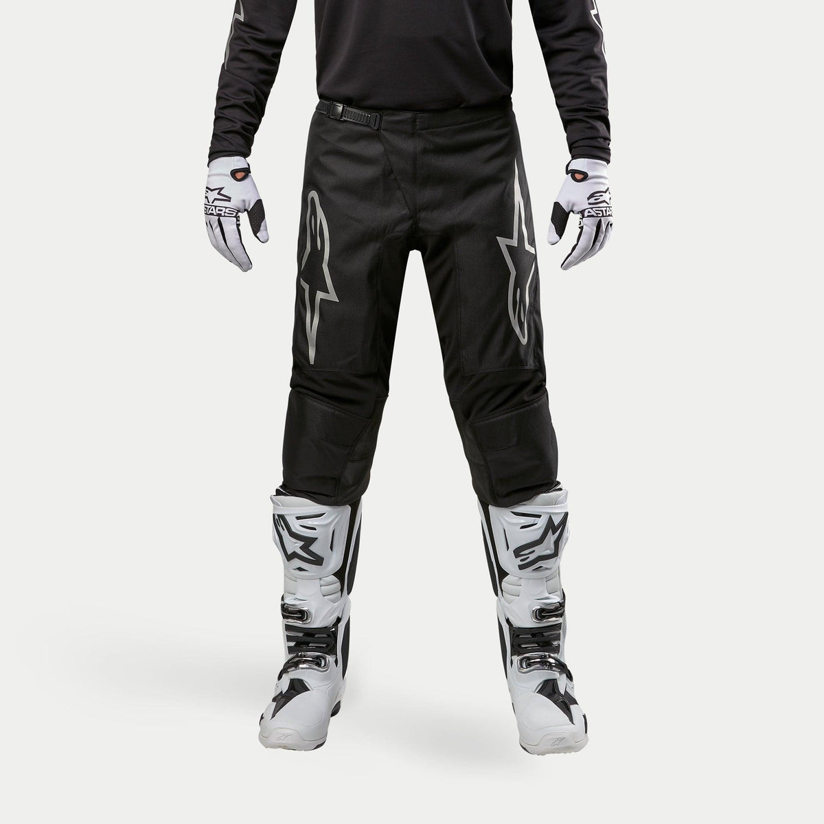 Alpinestars 2024 Fluid Graphite Pants, pantalones de motocross, negro y plata, con logotipos plateados de Alpinestars en los muslos, combinados con botas y guantes de motocross blancos y negros, diseño deportivo para competición off-road