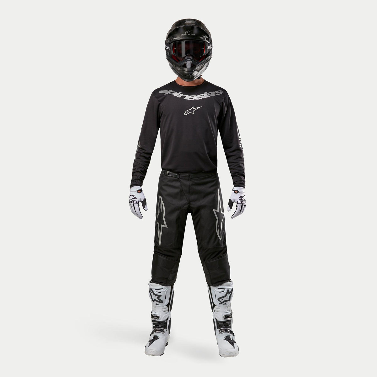 Piloto de motocross con equipamiento de carreras completo, pantalones Alpinestars 2024 Fluid Graphite en negro y plata, jersey negro de manga larga con logotipos plateados, casco negro con gafas, botas blancas y negras, guantes blancos y negros, conjunto coordinado para motocross