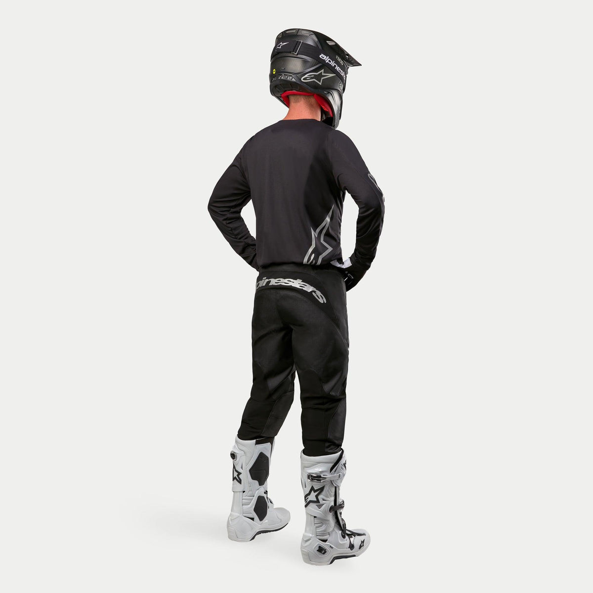 Pantalones Alpinestars 2024 Fluid Graphite, pantalones de motocross, negro y plateado, vista trasera de un piloto vistiendo jersey negro, casco negro y botas blancas y negras, con logotipos plateados en la parte inferior de la espalda y construcción de poli-tejido duradero para carreras todoterreno