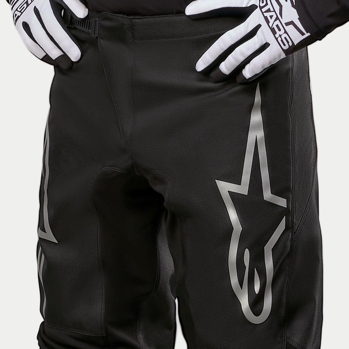 Alpinestars 2024 Fluid Graphite Pants, pantalones de motocross, negro y plateado, toma de detalle de la parte superior de los muslos y la cintura, con un gran logotipo de estrella plateada en la pierna, usados con guantes de carreras en blanco y negro