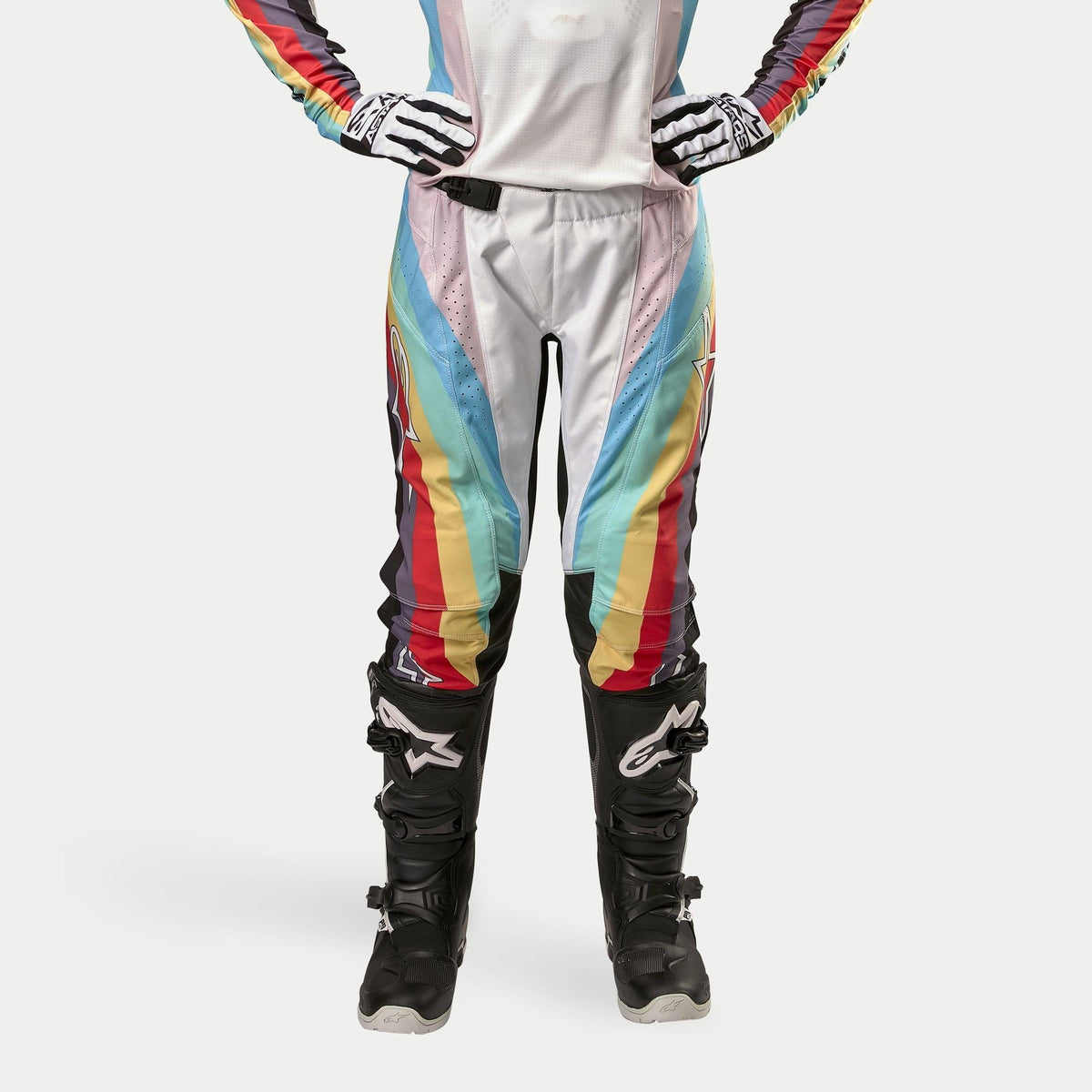 Alpinestars Stella Techstar Pants 2024 para mujer, pantalones de motocross, negro multicolor con vibrantes rayas diagonales en blanco, azul, rojo y amarillo, usados con botas y guantes de motocross negros, diseño inspirado en las carreras para rendimiento todoterreno