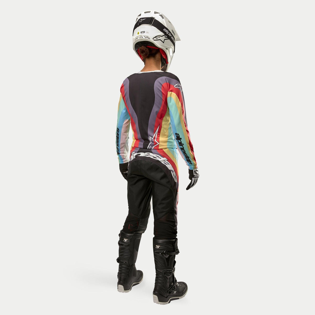 Corredora de motocross vista desde atrás vistiendo los pantalones Alpinestars Stella Techstar 2024 para mujer en Negro Multicolor, con un gran logotipo blanco en el asiento, combinados con un jersey de rayas de colores, casco blanco y botas Tech 3 negras para carreras todoterreno.